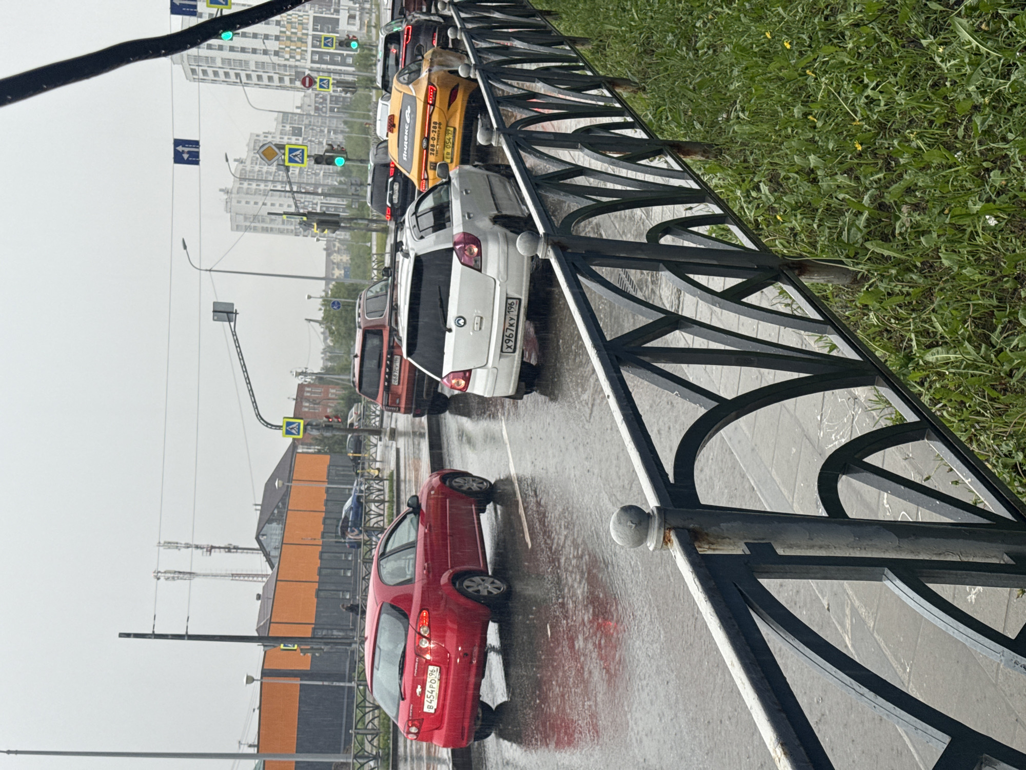 The image captures a street scene in Yekaterinburg, Russia, during what appears to be a rainy or recently rained-out day. The road surface is wet, showing reflections, and the sky is overcast.
The primary subjects are numerous cars stopped in traffic, a black metal fence running along the roadside, and urban infrastructure. In the foreground, a red hatchback (license plate B454PO 96) is parked or stopped. Further along the road, several cars are queued, including a white SUV (license plate X967KY 196) and a yellow taxi. The yellow taxi prominently displays "Яндекс.Такси" (Yandex.Taxi) branding and a phone number "288-0-288", along with its license plate EB 054 96. Other vehicles, mostly dark-colored, are visible in the distance.
The scene depicts a typical city street with multiple lanes. To the right of the road, a grassy verge with some dandelions separates the pavement from the fence. On the left side, modern high-rise apartment buildings are visible in the background, along with streetlights, traffic lights (some showing green), and road signs, including pedestrian crossing signs and directional arrows. The fence itself has decorative, arched sections. While no people are distinctly visible, their presence is implied by the vehicles and urban setting. The overall mood conveyed is that of a bustling city experiencing inclement weather.