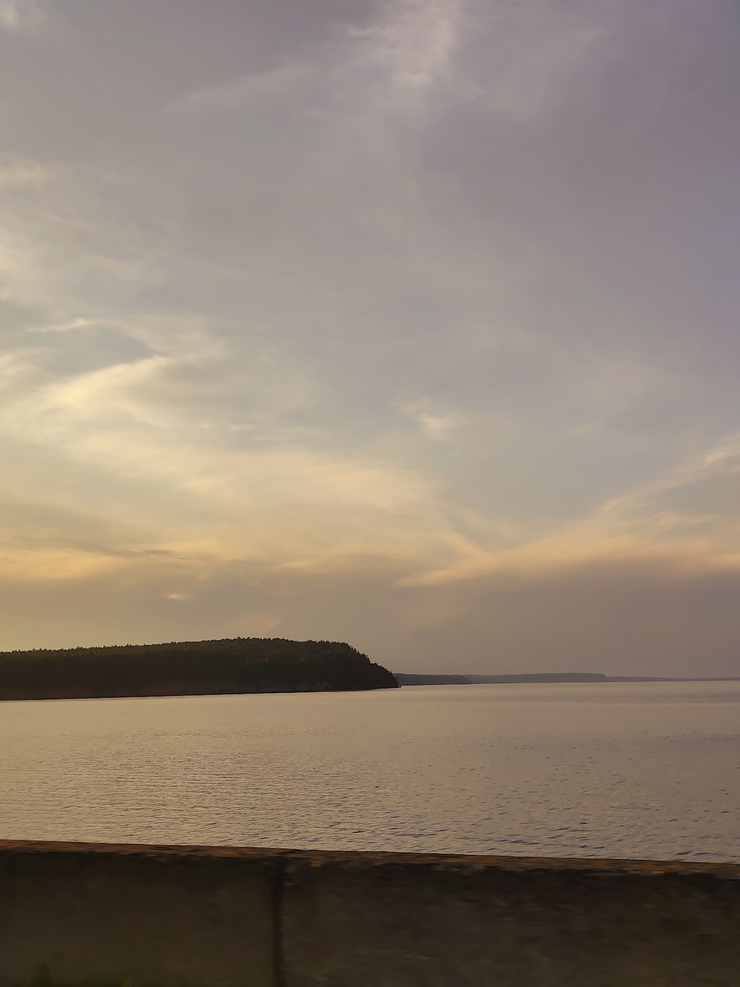 This image depicts a serene waterscape near Chaikovsky, Russia. A broad, calm body of water, likely the Kama River, occupies the lower half of the frame. The water's surface reflects the muted, warm tones of the sky, showing subtle ripples. Along the horizon, a dark, tree-covered shoreline stretches across the left and center of the image, forming a peninsula. The sky above is a canvas of soft, hazy clouds, tinged with hues of lavender, pale yellow, and peach, indicative of either dawn or dusk. In the foreground, a concrete barrier runs horizontally across the bottom of the image, partially obscuring the view of the water and suggesting a viewpoint from a road or embankment. There are no people or immediate human activity visible in this tranquil natural setting.