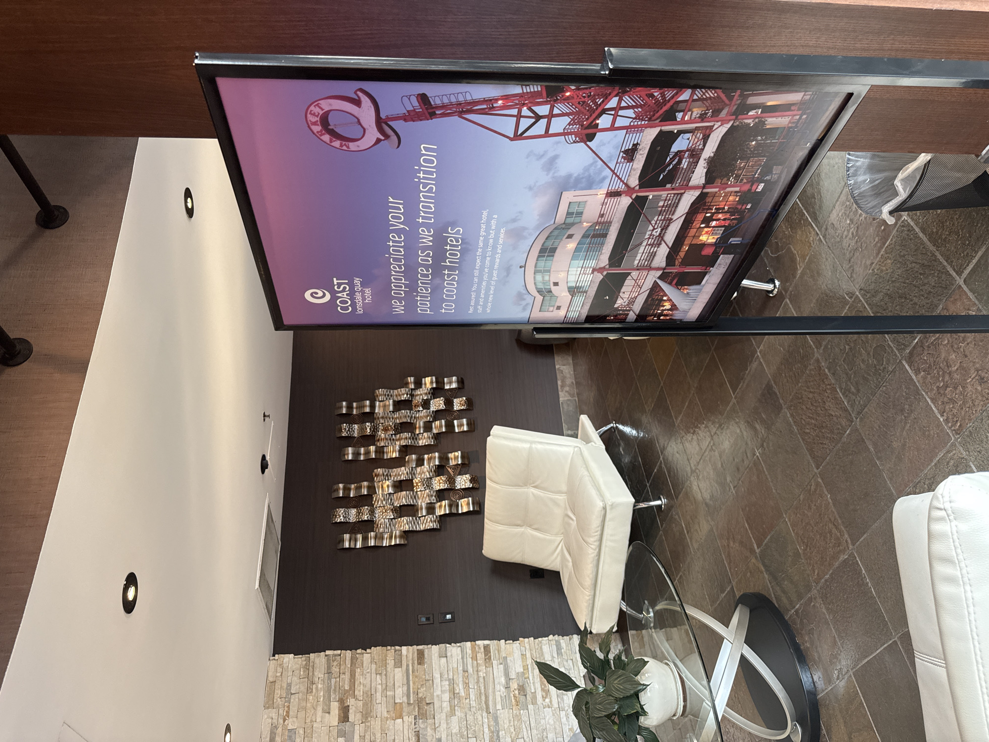 The image depicts the lobby of the Coast Lonsdale Quay Hotel in North Vancouver, Canada. A digital sign stands prominently in the foreground, displaying a message of appreciation for patience during a transition to Coast Hotels, accompanied by a photograph of the Lonsdale Quay Market. In the background, a modern, cream-colored sofa sits next to a glass coffee table with a plant. The walls are decorated with textured dark brown wallpaper and a striking metal art piece. The floor is covered in large, dark slate tiles.