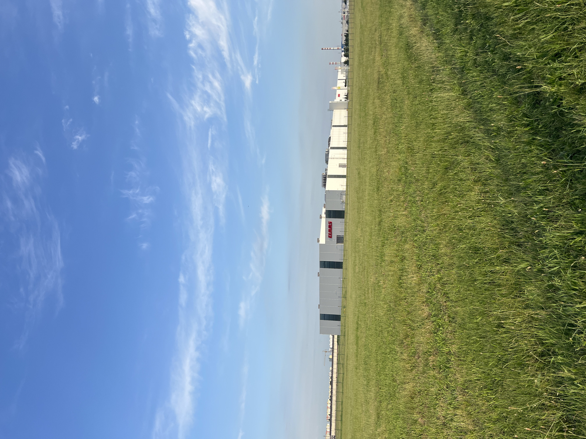 A long, white industrial building with red lettering that spells "CLAAS" is situated in the distance, behind a fence. The building is surrounded by a flat, grassy field under a clear blue sky with scattered white clouds. The field is covered in tall, green grass, and there are some industrial structures and a smokestack visible in the far distance. The scene appears to be located in a rural or industrial area.