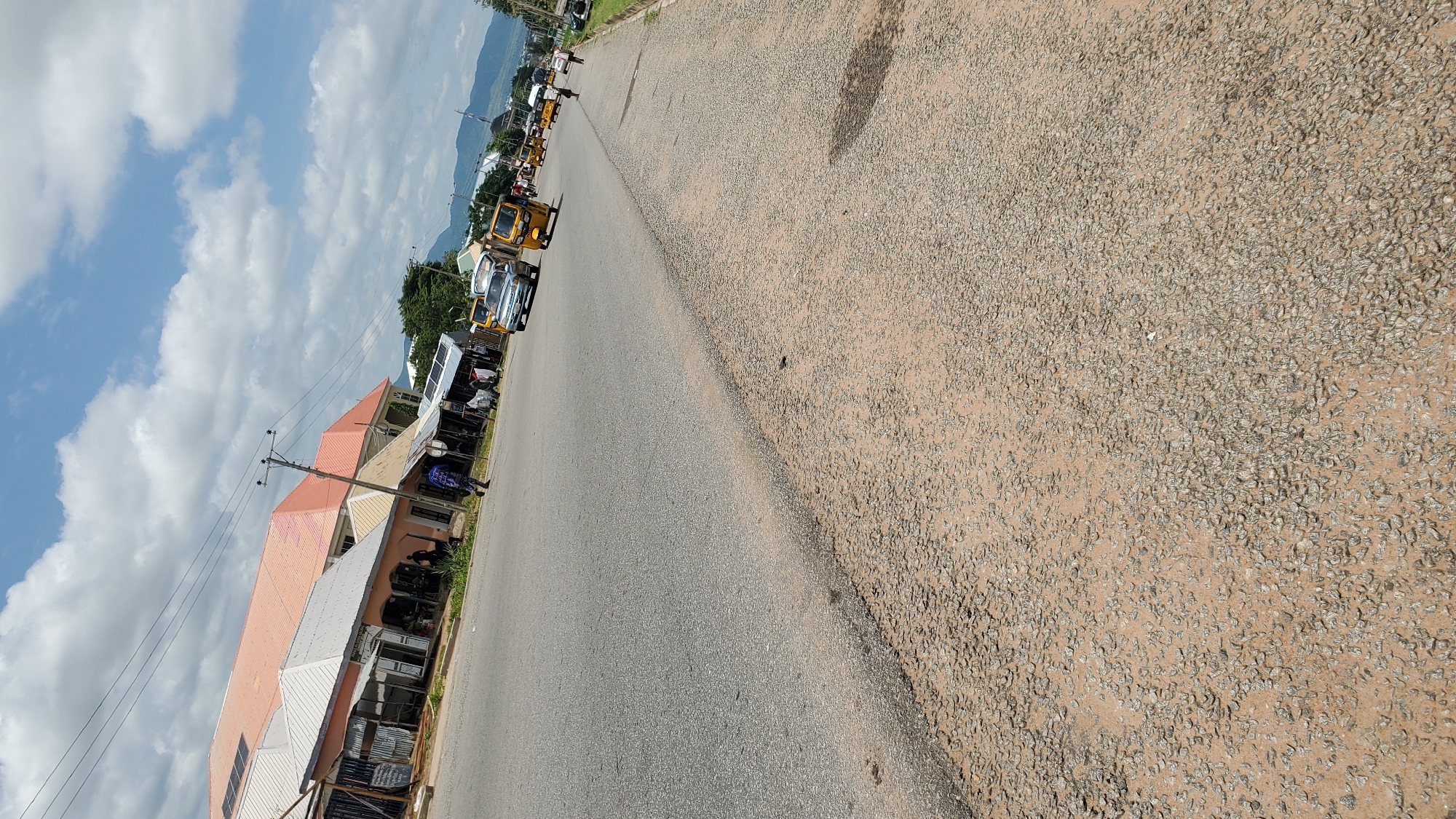 The image shows a street scene in Jalingo, Nigeria, with a paved road in the foreground and buildings and vehicles on the left side. The road appears to be a main thoroughfare with multiple lanes. On the left, there are several low-rise buildings with corrugated metal roofs and some with orange or pink roofing tiles. Some of the buildings have shop fronts. Several people are visible walking on the sidewalk or near the buildings. A few vehicles are on the road, including a silver car, a yellow auto-rickshaw (Keke Napep), and other vehicles further in the distance. Power lines are visible above the buildings. The sky is partly cloudy with blue sky and white clouds. The ground in the immediate foreground is unpaved and covered with gravel and sand. In the background, there are hills or mountains.