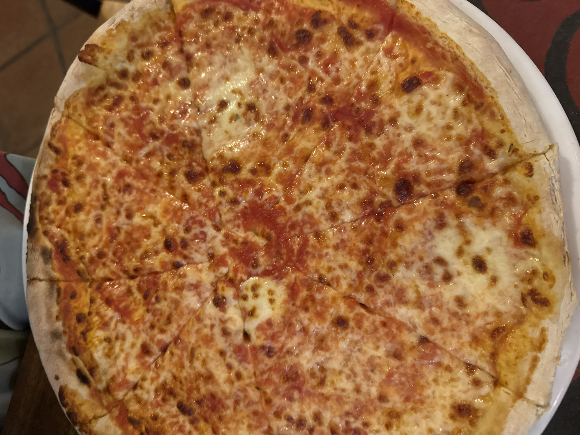 A round pizza, cut into slices, is presented on a white plate. The pizza appears to be a classic cheese pizza with tomato sauce and mozzarella cheese that is bubbly and slightly browned. The crust is thick and slightly charred. The image was taken in the city of Ungasan, Indonesia.