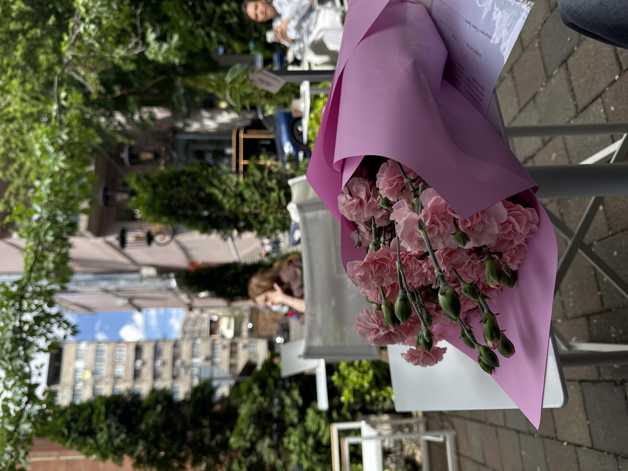 The image shows a bouquet of pink carnations wrapped in pink paper, hanging on a grey metal chair. A white piece of paper is partially visible behind the flowers. The scene appears to be an outdoor cafe or patio setting. In the background, other tables and chairs are visible. People are seated at some tables. Trees and buildings are also visible. Based on these visual cues, the location appears to be Yerevan, Armenia.