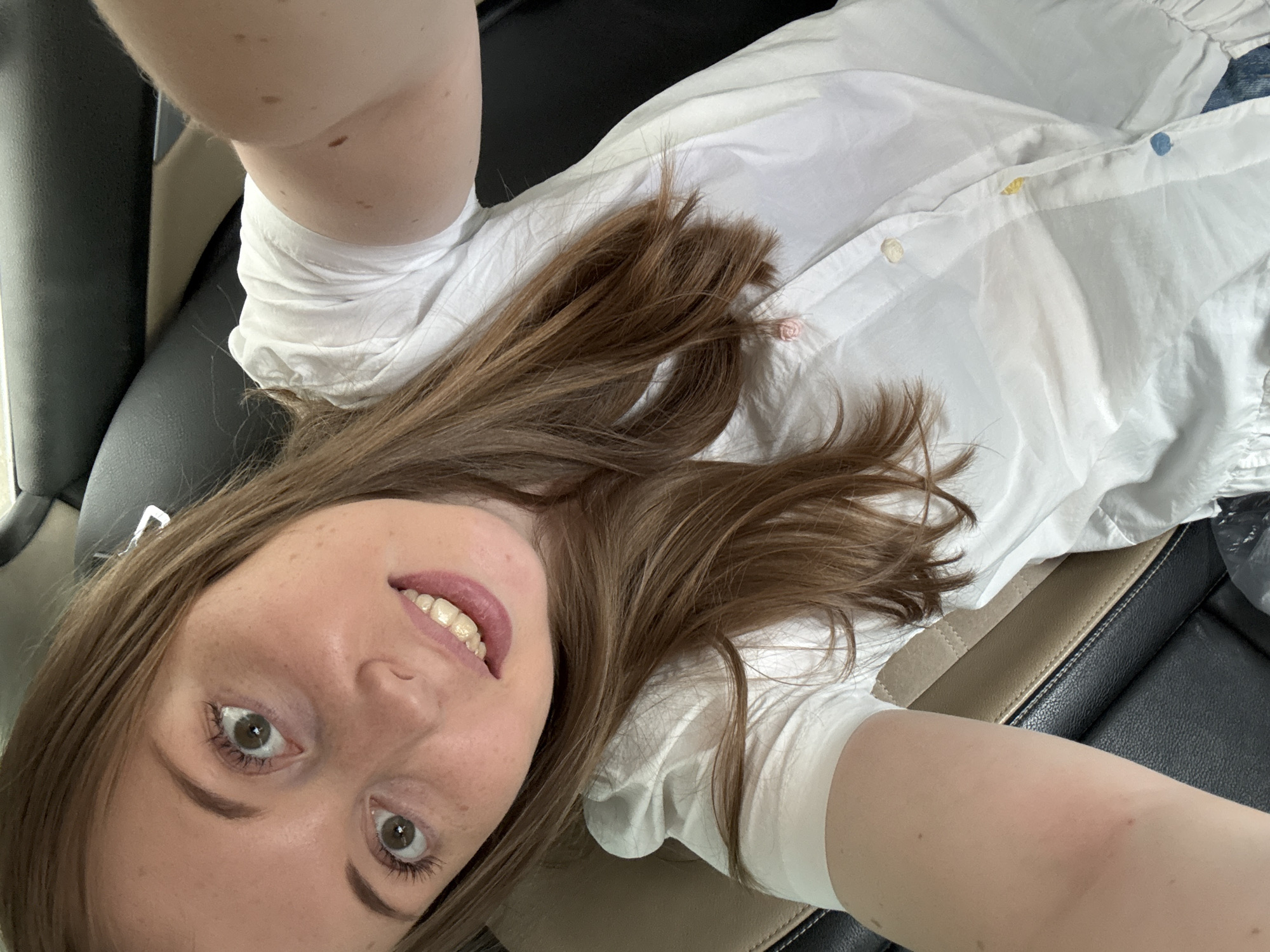 A young woman with long, brown hair and fair skin is taking a selfie inside a vehicle. She is wearing a white shirt with ruffled sleeves and colorful buttons. Her head is tilted, and she is smiling, showing her teeth. The background shows a car interior with black and tan seats. There is no visible indication of the specific location within the city.