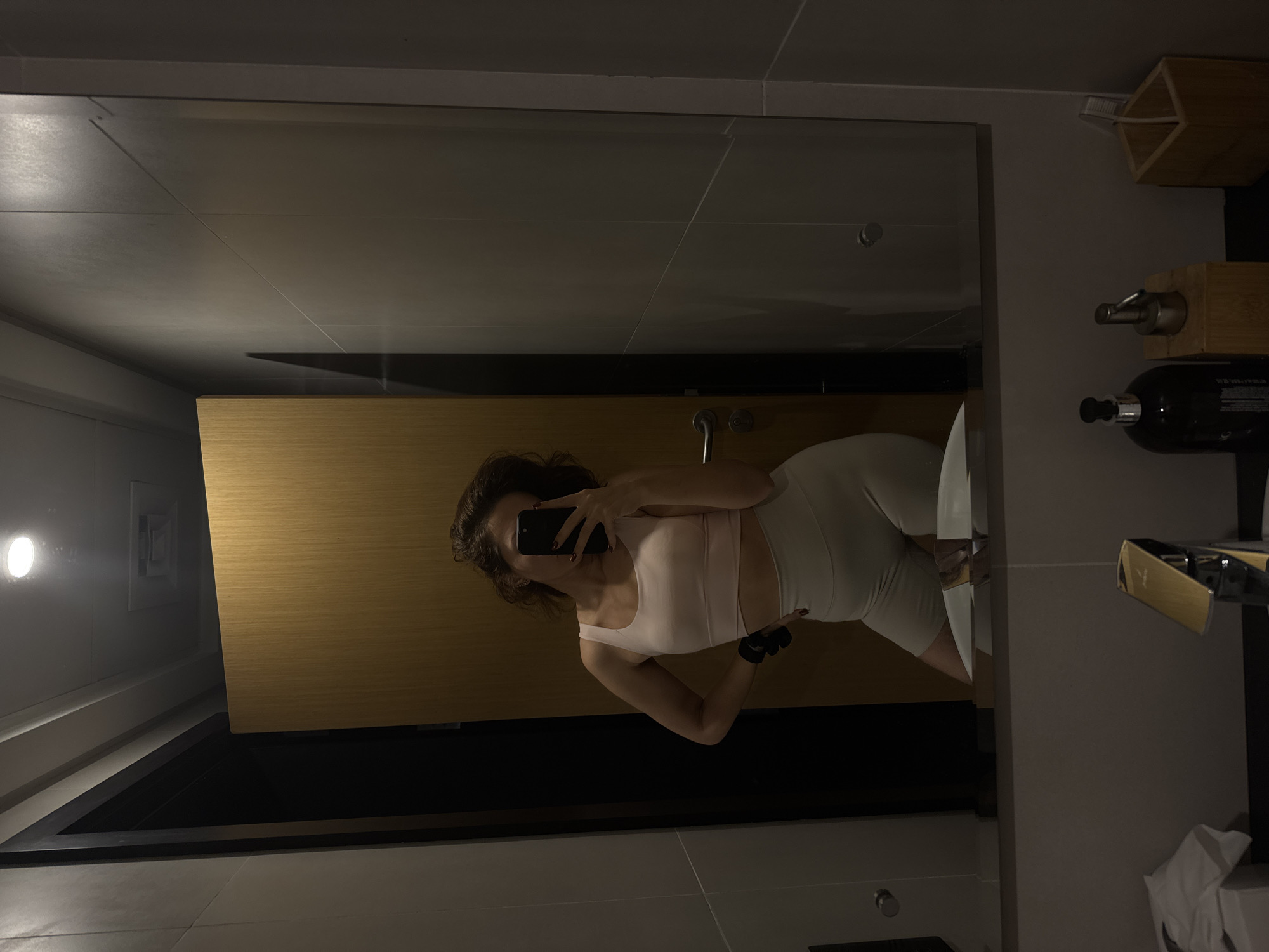 The image is a selfie taken in a bathroom. A woman with curly hair is visible in the mirror, wearing a light pink sports bra and light-colored shorts. She is holding a smartphone and taking the picture. Behind her is a wall with a wooden panel and a mirror. To the right of the mirror, there are bathroom accessories, including a soap dispenser and a wooden holder. The bathroom appears to be modern and well-lit. The location is Dubai, United Arab Emirates.