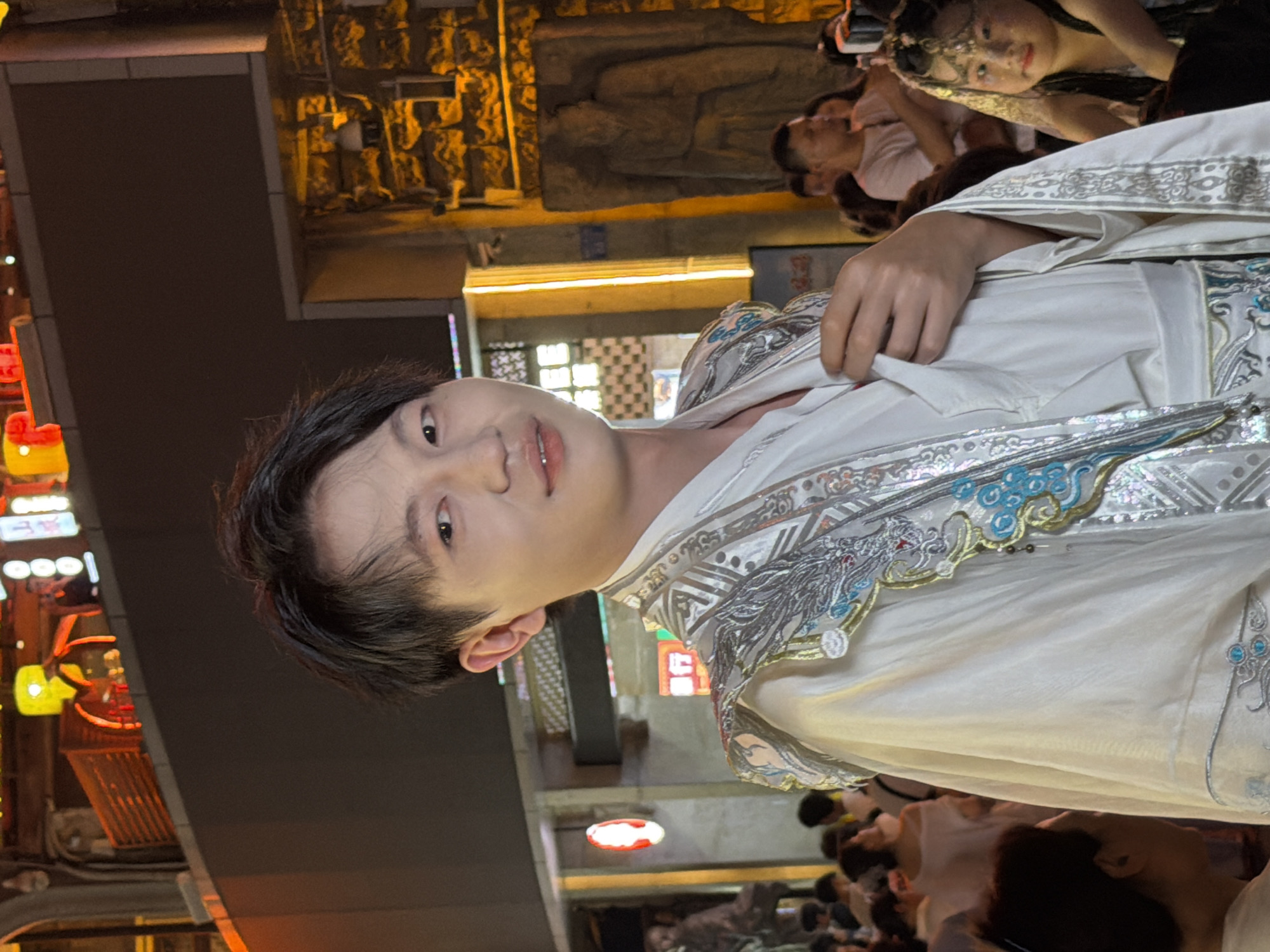 This is a photograph taken in Yuzhong District, China, likely at night given the artificial lighting. The scene depicts a group of people gathered, possibly for a performance or cultural event. The foreground is dominated by a young person dressed in an ornate, white garment with intricate silver and blue embroidery. This individual is looking towards the camera with a neutral expression.
Behind the main subject, other individuals are visible, some appearing to be performers or participants in the event. One woman in the background wears a delicate headdress. The setting suggests an outdoor or semi-outdoor area with architectural elements and illuminated signage, hinting at a commercial or entertainment district. The lighting is warm and inviting, casting a glow on the surroundings. The overall atmosphere appears festive and engaging. No legible text is visible in the image.