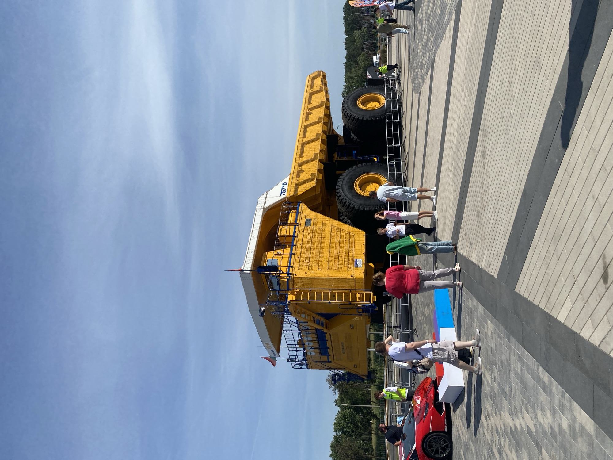 A large, bright yellow mining truck, identified by the "BELAZ 75710" marking, is the central subject of the image. The truck has massive black tires and a white cab. Several people are gathered around the truck, some standing and observing, while others appear to be walking. In the foreground to the left, a red sports car is visible. The scene is set outdoors on a paved area with a clear blue sky overhead, suggesting it is daytime. The background consists of trees and some distant structures, hinting at an urban or semi-urban environment. The location is identified as Minsk, Belarus.