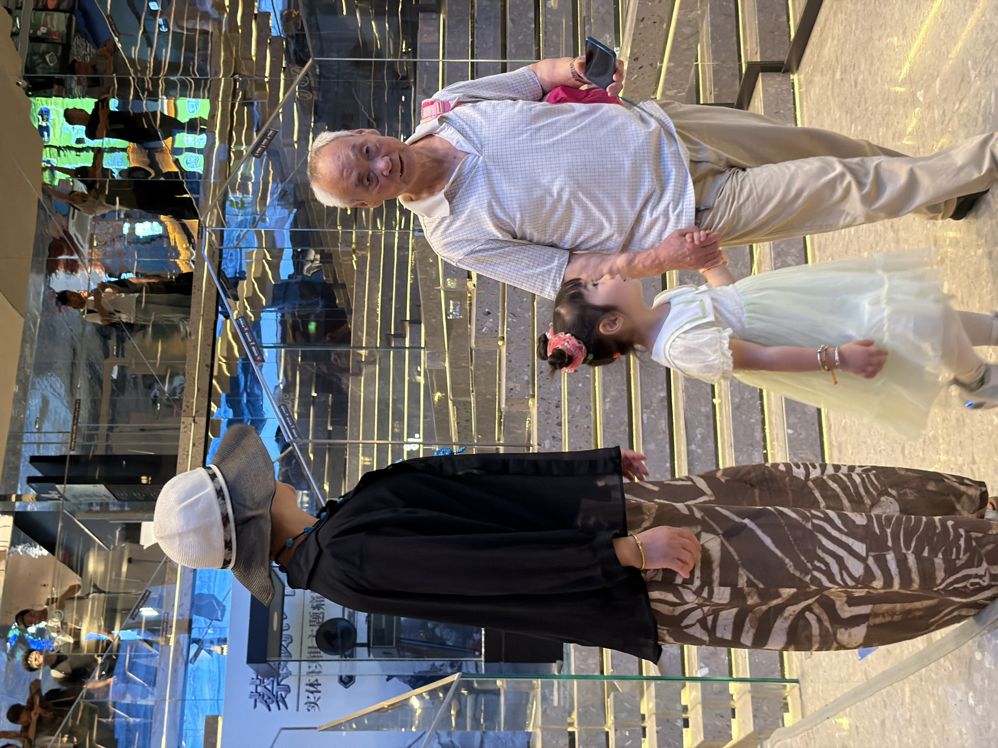In Yuzhong District, China, an elderly man, a woman, and a young girl are walking together. The man, on the right, wears a light-colored plaid polo shirt and beige pants, and carries a pink bag over his shoulder. He holds the girl's hand. The girl, in the center, wears a light green dress with puffed sleeves and has a pink hair accessory. The woman, on the left, wears a black shirt, a wide-brimmed white and gray hat, and patterned brown pants. She also wears a turquoise necklace. They are walking on a polished floor with reflective walls and a staircase in the background, suggesting an indoor, possibly commercial or public, location.