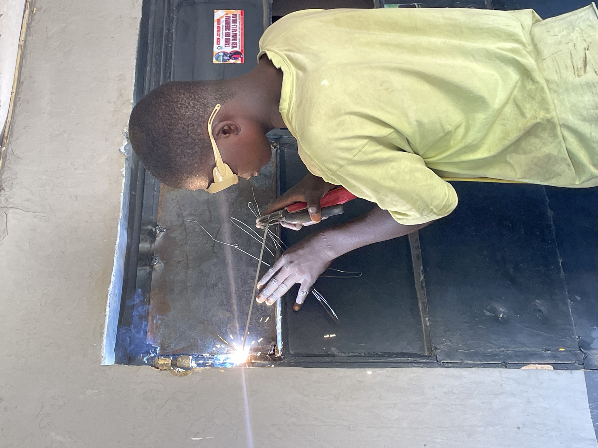 A person is welding a dark metal frame inside a light gray doorway. The person wears a light yellow shirt and protective eyewear. Sparks are visible from the welding site. A sign with text in a local language and images is affixed to the door frame above the person. The location is inferred as Jos, Nigeria, based on the text on the sign.