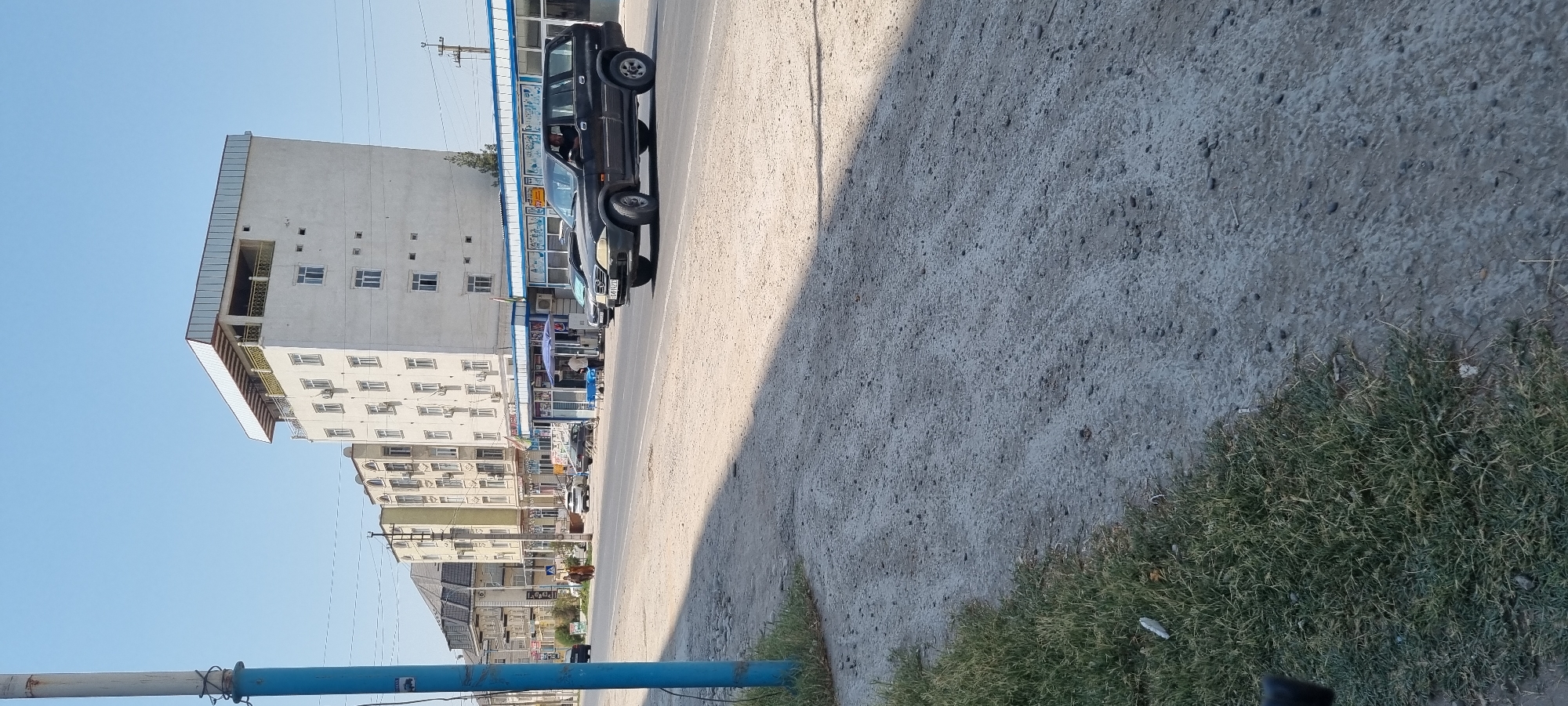 In Ismoili Somoni, Tajikistan, a street scene unfolds under a clear blue sky. A dark-colored SUV is captured in motion on the paved road, its front left wheel slightly turned. The vehicle appears to be a SsangYong Rexton, based on its distinctive design. A person is visible in the driver's seat. To the right of the SUV, a black sedan is also on the road. The background features a row of buildings with multiple stories, some displaying signs. One building, a tall white structure, has a distinctive metallic roof with an overhang and a balcony. In the foreground, a dirt and gravel shoulder lines the road, with patches of grass on the left side. A blue pole stands on the far left, partially obscuring the view of the buildings. The street is casting a long shadow from the right, indicating the time of day is likely late afternoon or early morning.
