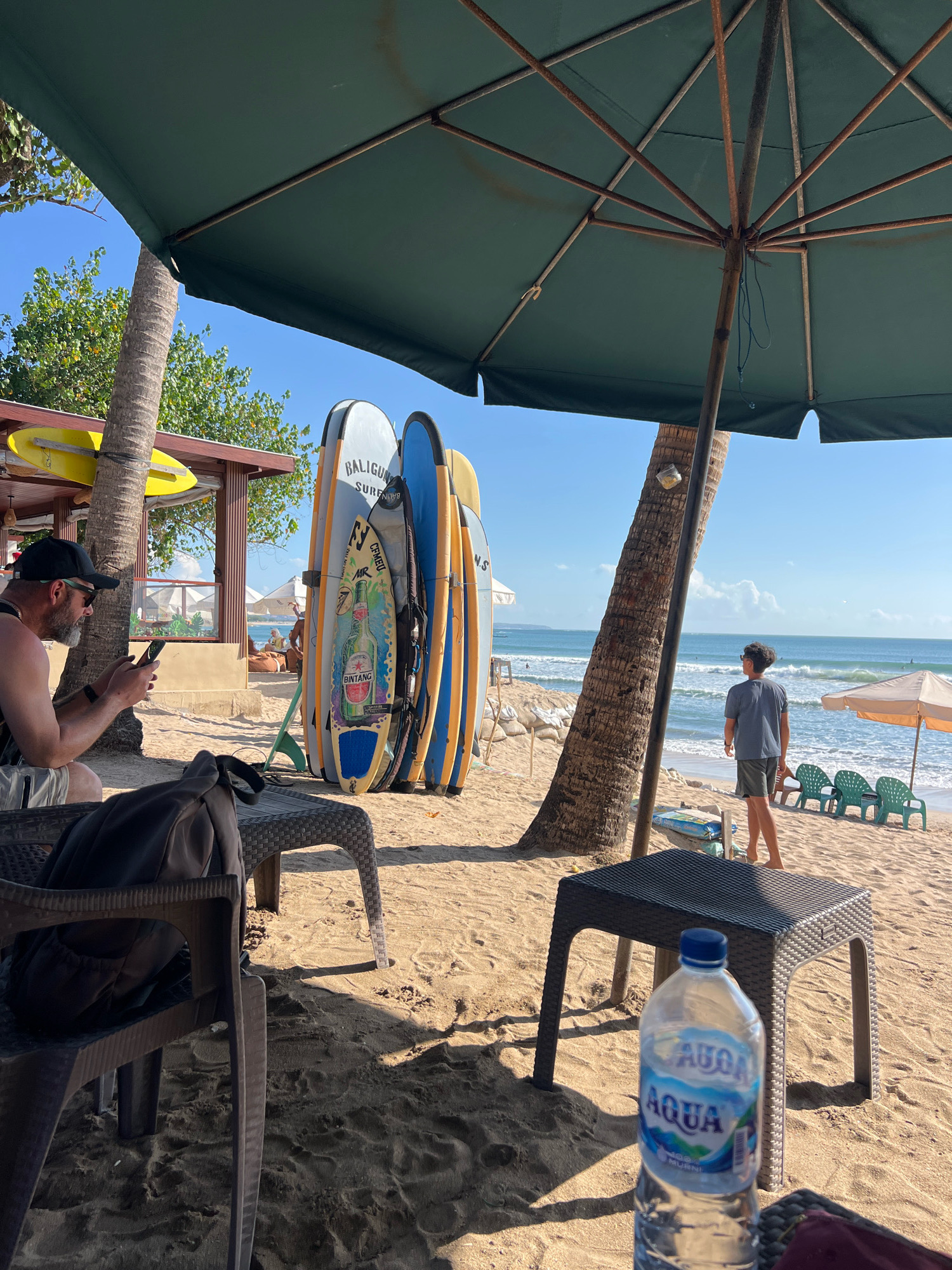 This image depicts a beach scene in Kuta, Indonesia. In the foreground, a man wearing a black baseball cap and sunglasses is seated on a chair and looking at his phone. A black bag is placed on a woven plastic table next to him. A bottle of Aqua water is on another table in the right foreground. Behind him, several surfboards are propped up, leaning against a palm tree. In the mid-ground, a young person in a grey t-shirt and shorts stands facing the ocean. The beach has fine sand, and the ocean has moderate waves. Several beach chairs and umbrellas are visible in the distance. The sky is clear and blue, indicating a sunny day. The overall atmosphere is relaxed and typical of a beach resort.