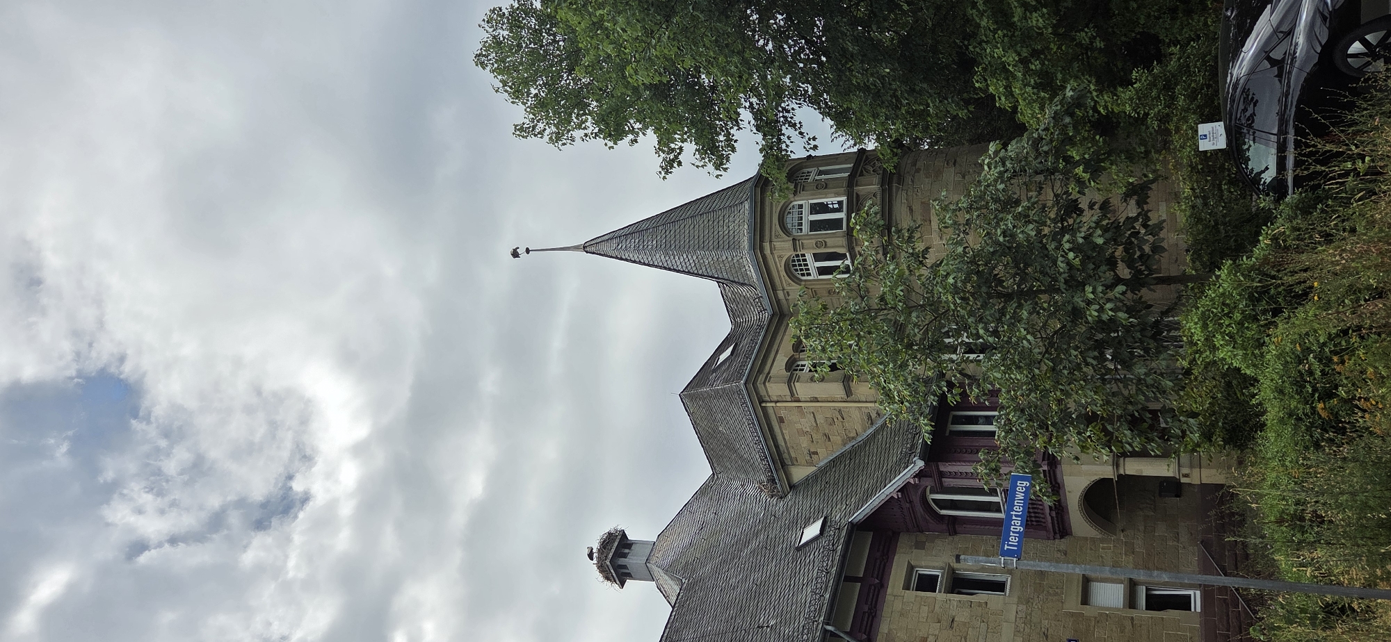 The image shows a historical building with a slate roof and stone facade, featuring a distinctive turret with a weather vane. A stork's nest is visible on a chimney on the left side of the building, with at least one stork perched in it. A street sign reading "Tiergartenweg" is mounted on a pole in front of the building. The building is surrounded by lush green trees and vegetation. A black car is parked in the foreground, partially obscured by bushes. The sky is overcast with dramatic clouds. This building is likely located in Karlsruhe, Germany, given the street name "Tiergartenweg," which is a known street in the city.