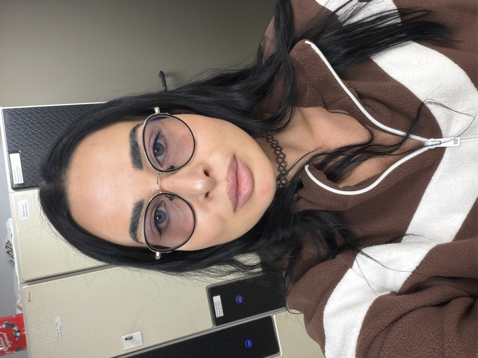 A medium close-up, vertically oriented image (originally rotated 90 degrees clockwise) features a young woman posing for a photo. She has long, dark, wavy hair and is wearing round, metal-rimmed glasses with slightly tinted lenses. Her blue eyes look directly at the viewer, and her lips are softly parted. She wears a black choker necklace and a brown fleece jacket with wide white stripes and a white zipper running down the front.
The background consists of light-colored, modular storage or filing cabinets, possibly metal, arranged vertically. Several black panels are visible on these cabinets, containing white numbers such as "452" and "129." Small, white labels with black Cyrillic text are also affixed to the cabinets. Visible text snippets include "Сведения ВУЗА" (Information about the University/Higher Education Institution), "Пример УП и ГЗ" (Example of Educational Plan and State Exam), and a partially visible "Офисный короб" (Office box) on a small red item in the lower-left corner. These textual cues suggest an academic or administrative environment, such as a university office or archive, in Saint Petersburg, Russia. The lighting is even, suggesting an indoor setting without strong natural light cues. The woman maintains a neutral to slightly pensive expression.