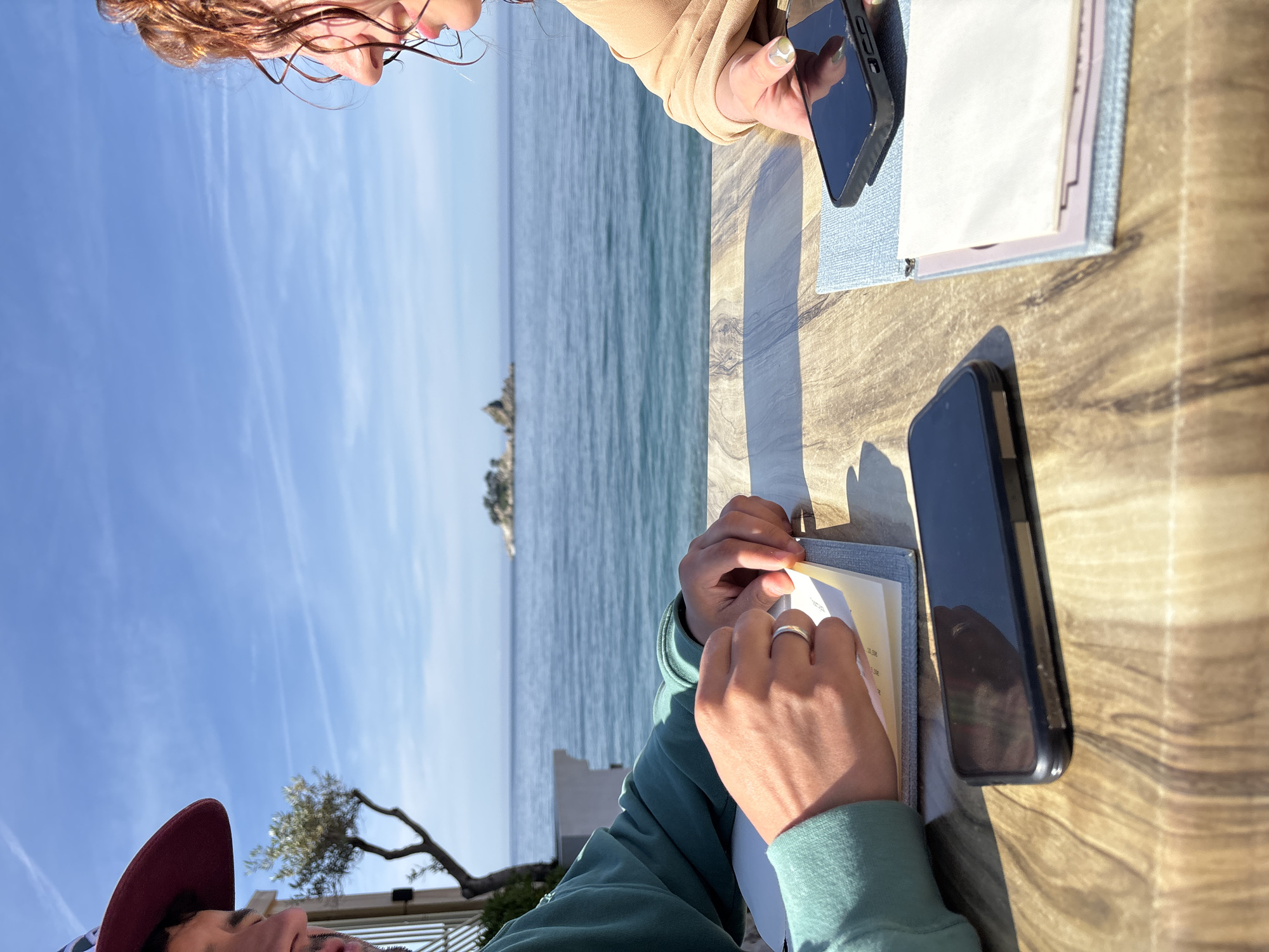 The image shows two people seated at a wooden table in Petrovac centar, Montenegro, based on the seascape visible in the background. One person, wearing a tan top, holds a smartphone in their right hand next to a light-colored menu. The other person, wearing a green sweatshirt and a burgundy hat, is looking down at a similar menu. A second smartphone is lying on the table near them. The sea is visible in the background, with an island or rock formation in the distance under a blue sky.