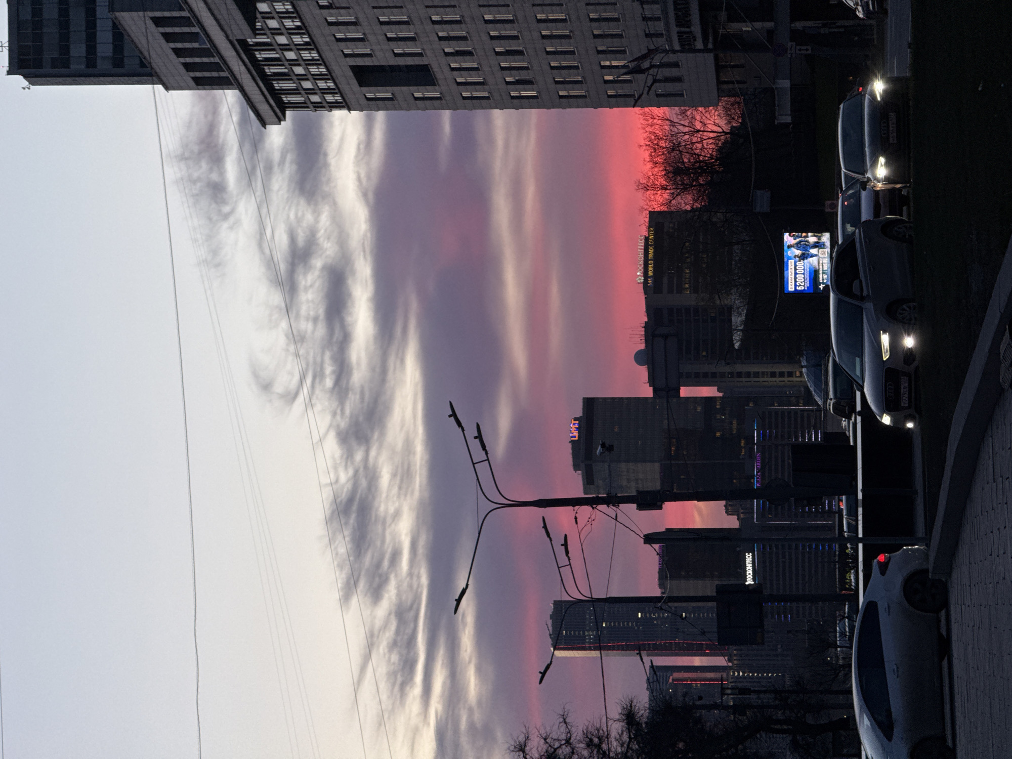 The image depicts a street in Moscow, Russia, at dusk. A tall building is visible on the left side of the frame, with the sky transitioning from light grey to vibrant pink and purple hues. A series of modern buildings and skyscrapers are visible in the background, silhouetted against the colorful sky. Streetlights stand along the street, with utility lines running across the sky. Several cars are parked along the right side of the street, their headlights illuminating the scene. An advertising billboard is also visible on the right side of the frame.