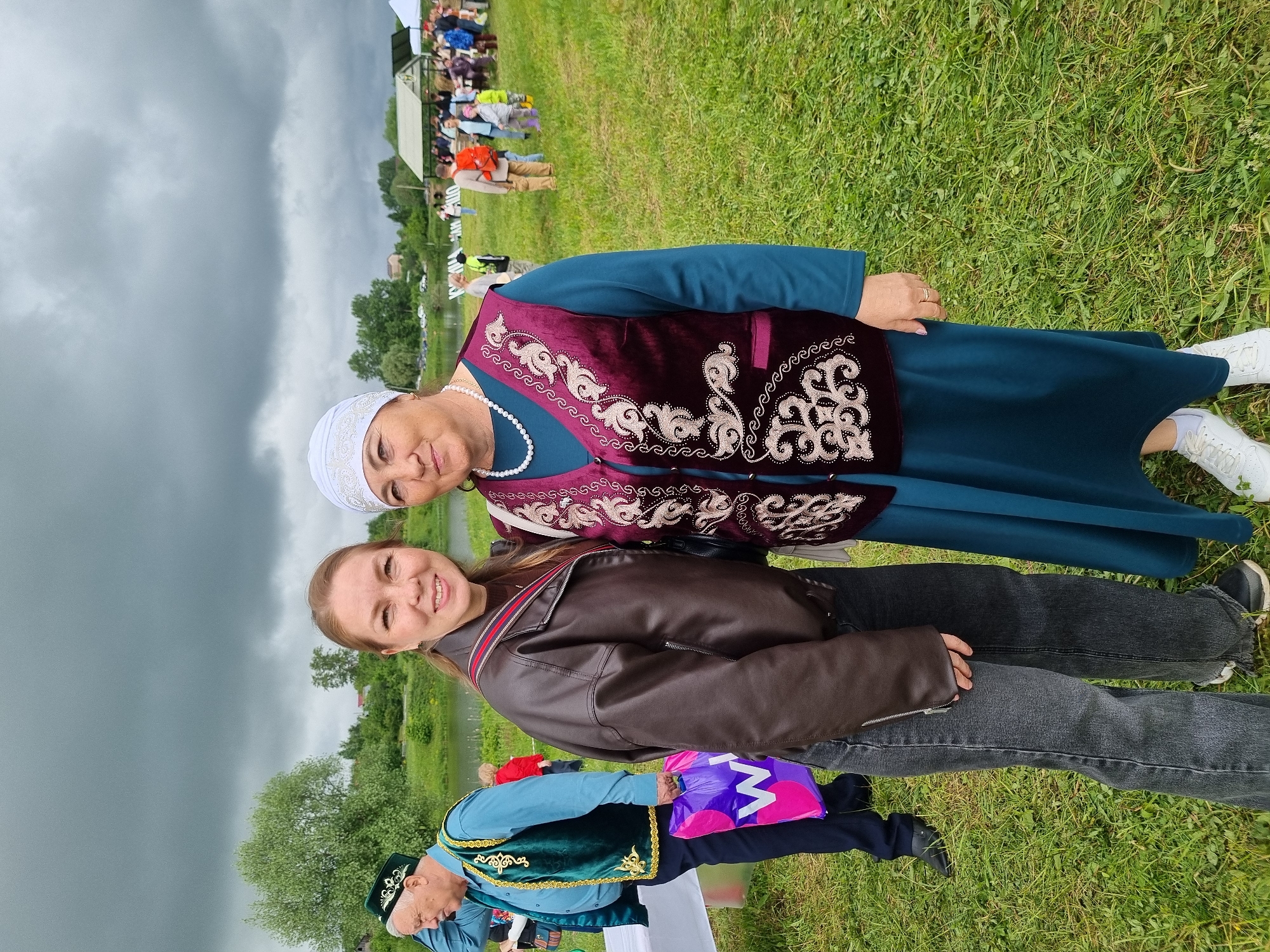 This image captures an outdoor scene in Фоки, Russia, on an overcast day. Two women are centrally positioned, posing for a photograph. The woman on the left, appearing to be in her late 20s or early 30s, has light brown hair pulled back and is smiling directly at the camera. She wears a dark brown leather bomber jacket over a dark top, dark gray jeans, and dark sneakers. The woman on the right, appearing to be in her 50s or 60s, is also smiling at the camera. She wears a white headscarf with an embroidered front edge, a long-sleeved teal dress, and a maroon velvet sleeveless vest intricately embroidered with gold or silver floral patterns. A white pearl necklace is visible around her neck, and she wears white sneakers.
In the left background, an older man stands near a white table. He wears a light blue shirt, dark pants, a dark green vest with gold trim and embroidery, and a matching green traditional cap. He holds a purple shopping bag with a white "W" logo and appears to be adjusting his cap.
The background features a grassy field with scattered groups of people, including adults and children, suggesting a public event. A man with a red backpack is visible in the mid-right background. Beyond the field, a body of water (river or pond) is visible, with green trees and foliage lining its far bank. Distant buildings or houses are visible on a slight rise. In the mid-background, a green-roofed structure, possibly a gazebo or tent, and other white tents are present. Partially visible white signage with dark letters, possibly Cyrillic, is also in the background. The sky is heavily overcast with dark gray clouds, providing diffused lighting to the scene.