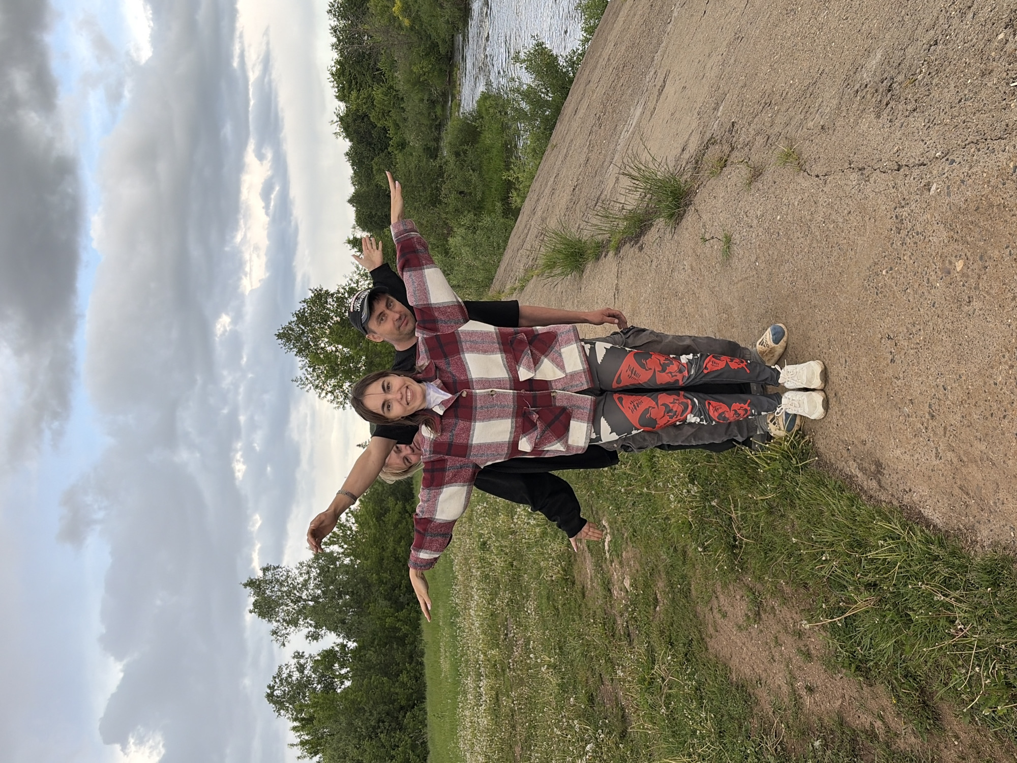 The image shows three people outdoors. A person wearing a baseball cap is standing behind a younger person wearing a red and white plaid shirt, which is covering another person. The three people are lined up and have their arms extended outward, resembling the shape of an airplane. They are standing on a grassy area next to a concrete structure. A river and wooded area are visible in the background. The sky is cloudy. The location appears to be in a park near a river in Nizhnii Vishur, Russia.