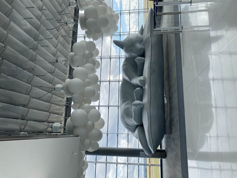 The image depicts the interior of a spacious terminal hall at Paris Charles de Gaulle Airport, Terminal 2E, located in Roissy-en-France, adjacent to Mitry-Mory. The primary subject is a large, light grey sculpture of a cat sleeping on its side atop a rectangular pillow, positioned centrally in the hall. Inscriptions "L'ESPRIT DU VENT" and "A. GILLAUD," along with Japanese text, are visible on the pillow. The ceiling features segmented, light grey paneling, from which numerous clusters of spherical, opaque white light fixtures, resembling bubbles, are suspended. A vast glass curtain wall forms the background, providing an unobstructed view of an airfield with green ground cover and distant infrastructure under a partly cloudy sky. The floor is composed of highly polished, light-colored tiles, exhibiting prominent reflections of the sculpture and overhead lighting. A dark grey cylindrical support pillar is visible on the far left, and a metal stanchion barrier is partially visible on the far right. No individuals are present in the scene.