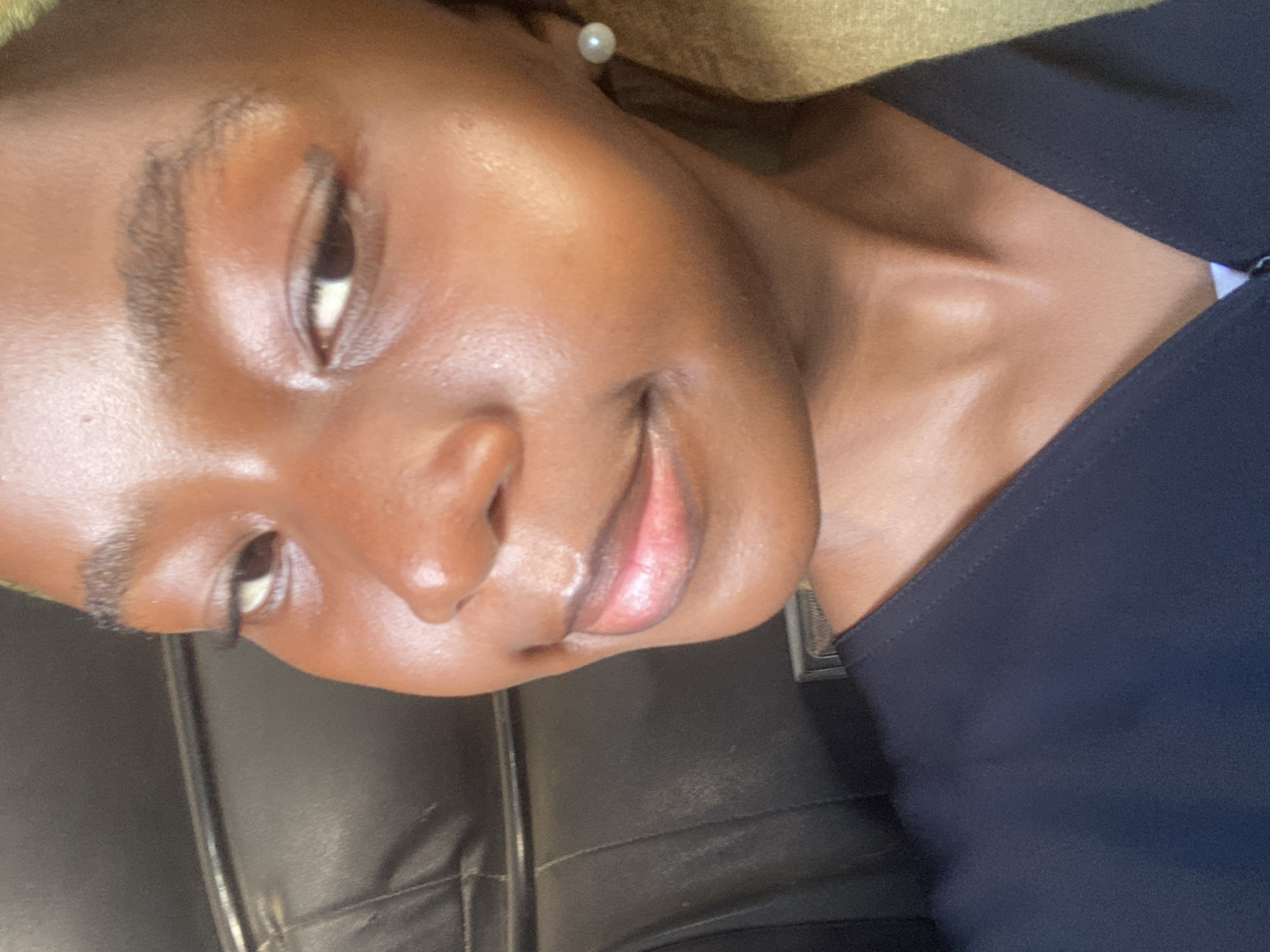 The image is a close-up, tilted portrait of a woman with brown skin. She has dark eyes, a small nose, and full lips. She's wearing a dark blue top and a pearl earring. The background is blurry, appearing to be a leather surface and a beige wall. There is no information available to identify this image as being located in the city of Dar es-Salaam, Tanzania.