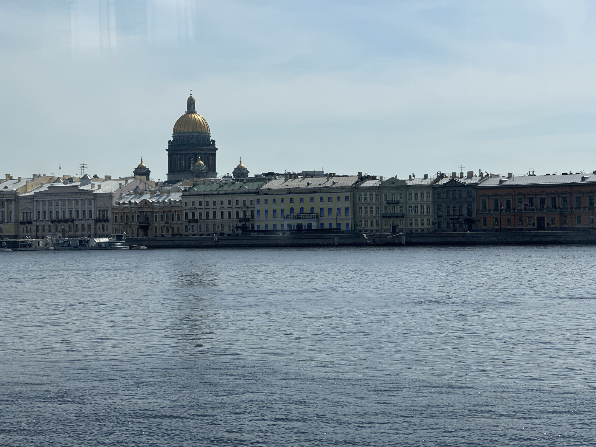 The image shows a cityscape across a body of water. In the background, a row of buildings lines the waterfront, displaying various architectural styles and colors, including white, yellow, and orange. A prominent golden dome, likely a church or cathedral, rises above the buildings. Boats are docked along the shore. The body of water in the foreground has a rippled surface, with reflections of the buildings and sky. The sky is overcast, with a pale blue hue.
The golden dome is that of Saint Isaac's Cathedral, placing the location in Saint Petersburg, Russia.