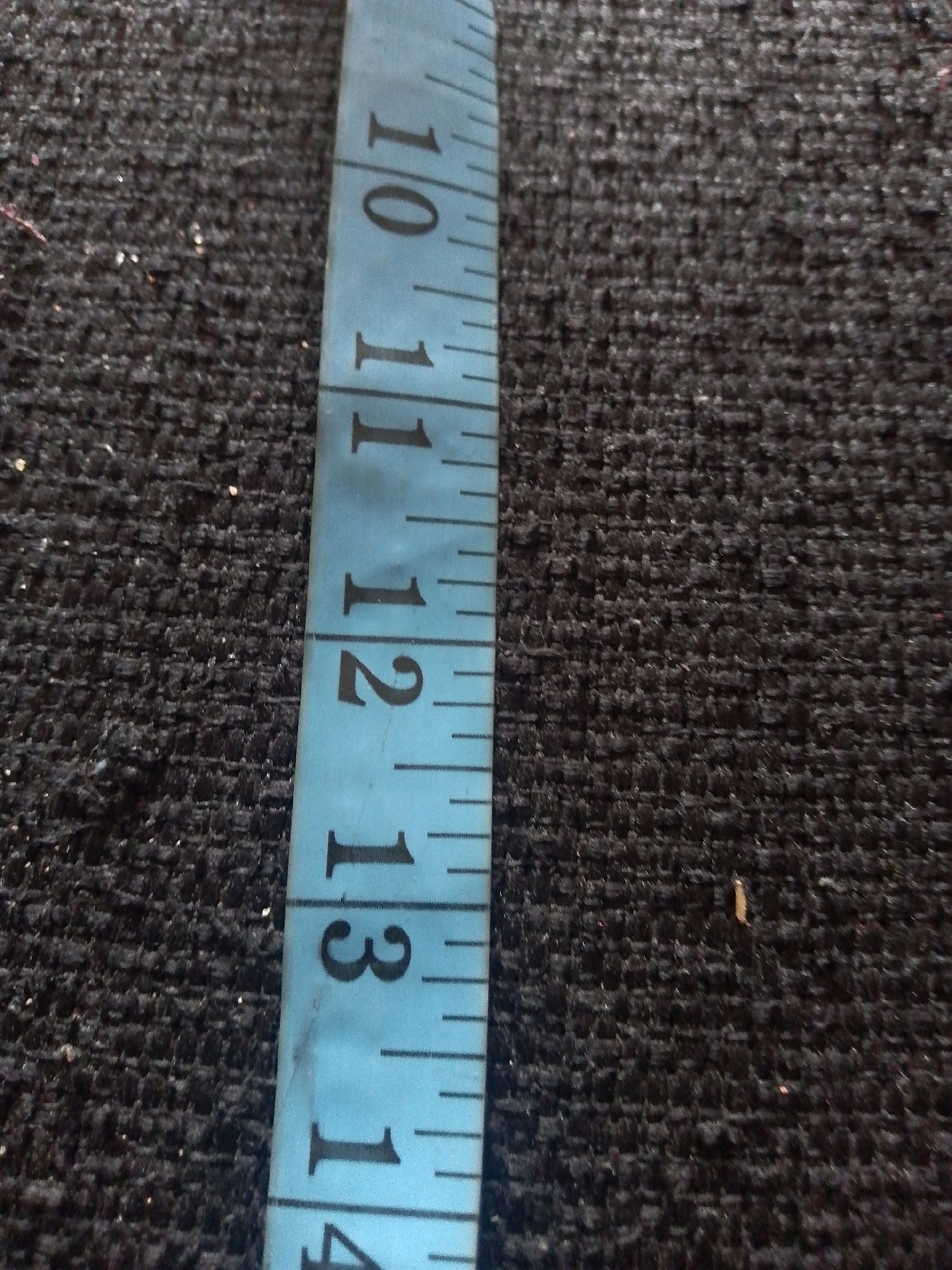 The image displays a close-up, angled view of a light blue measuring tape placed on a dark, textured fabric surface. The measuring tape is marked with black numerical indicators, including "10", "11", "12", and "13", along with smaller tick marks indicating increments. The fabric surface appears to be a woven material, possibly upholstery or carpet, with a rough texture. The lighting is consistent, with no strong shadows cast. The context of Jauro Yinu, Nigeria, is not visually represented in the image itself. There are no people, actions, or interactions depicted. The scene is static and focuses solely on the measuring tape and the fabric.