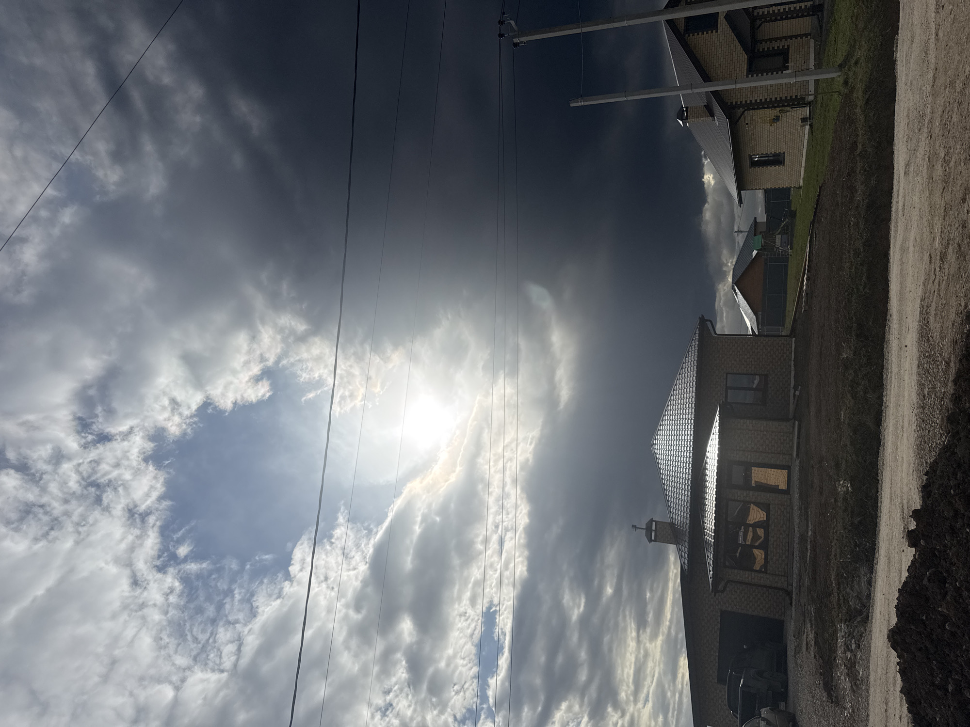 This image captures a rural or semi-rural landscape under a dramatic and dynamic sky. The scene is dominated by a mix of heavy, dark storm clouds on the left and upper parts of the frame, contrasting with brighter, whiter clouds on the right. The sun is intensely visible through an opening in the clouds in the upper right, casting a bright flare and illuminating specific areas.
In the mid-ground and foreground, several houses are visible. A two-story house on the right features light-colored brick walls and a dark, gabled roof with a chimney. Multiple windows reflect the bright sky. A dark vehicle is partially visible, parked in front of this house. Another house with similar construction is visible further left and in the background.
Multiple dark power lines stretch across the sky, running diagonally and vertically, supported by utility poles which are partly visible. A rough, unpaved dirt road or path runs along the bottom and right side of the foreground, with patches of dry grass and dirt surrounding the buildings.
The time of day appears to be daytime, likely late morning or afternoon, given the sun's position and intensity through the clouds. The weather is characterized by significant cloud cover with periods of bright sunlight. No specific activity or people are visible in the static scene. No visible text is present in the image. The setting, with its houses, dirt road, and power infrastructure, is consistent with a location such as Ферма, Russia.