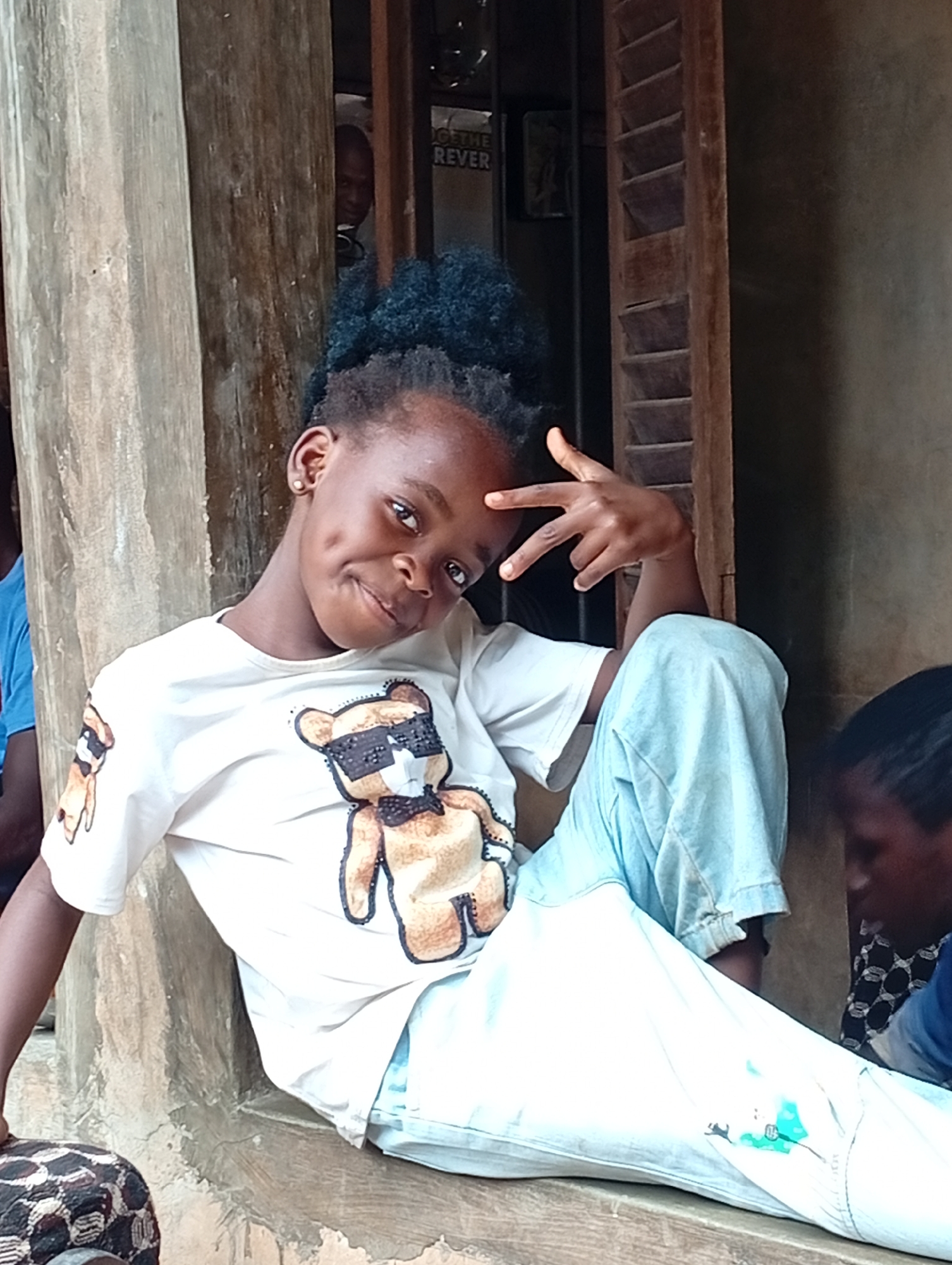 A young Black girl with dark curly hair and dimples is sitting on a ledge outdoors. She is wearing a white t-shirt with a graphic of a bear wearing sunglasses and a bow tie, and light blue jeans. She is striking a peace sign with her right hand. The background consists of a wooden structure, a doorway, and a wall. A person's head is visible in the doorway behind her. Another person is seated to the right of the girl, partially visible and facing away. The setting appears to be in Adda, Nigeria.