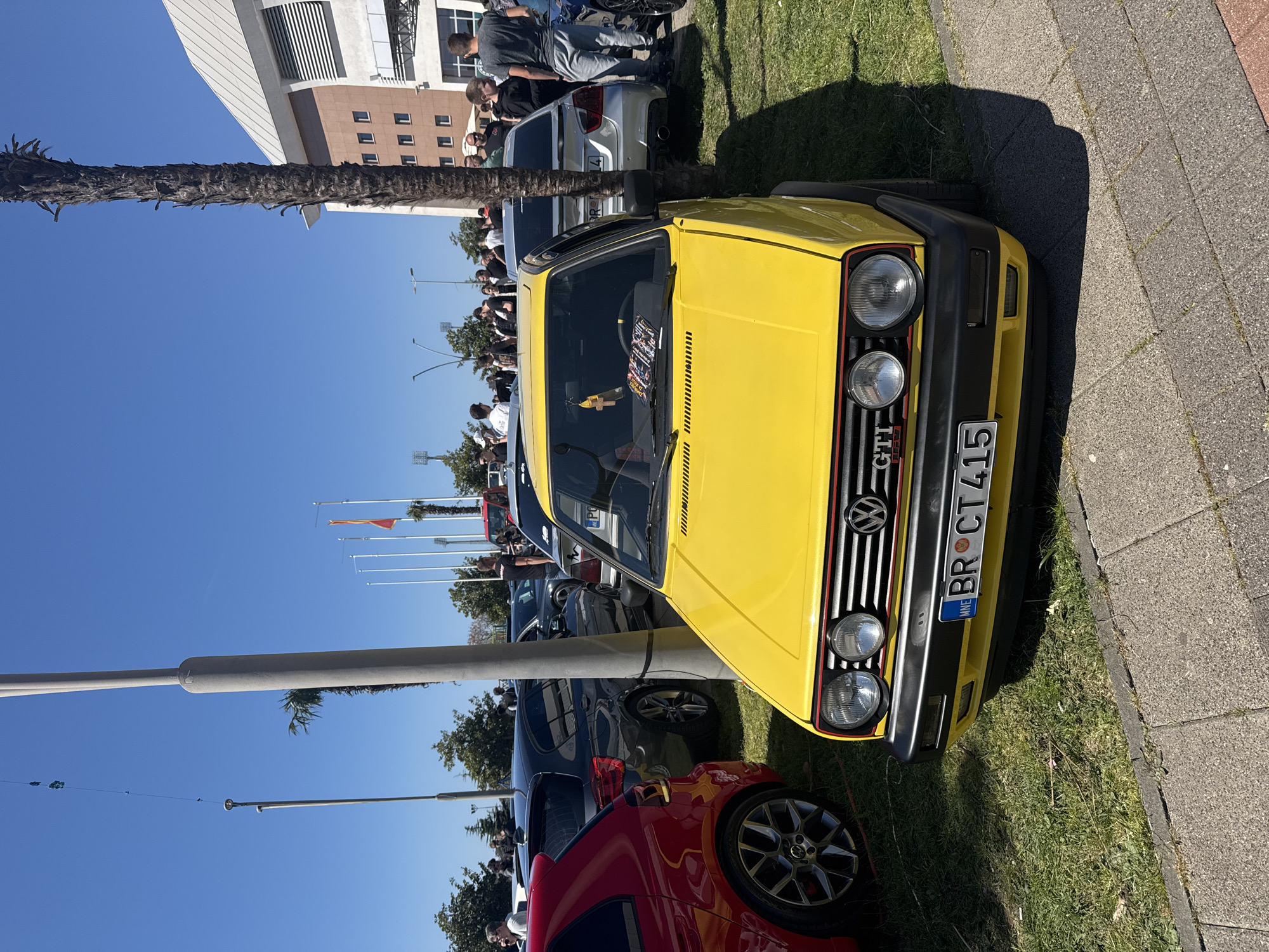 A bright yellow Volkswagen Golf GTI is parked on grass alongside a sidewalk in Bar, Montenegro. The car's license plate reads "BR CT 415". Several other cars, including a red hatchback and a silver sedan, are parked nearby. A crowd of people is gathered behind the cars, suggesting a car show or event. A palm tree and flagpole are in the background. The sky is clear and blue. A building with a modern design is also visible.