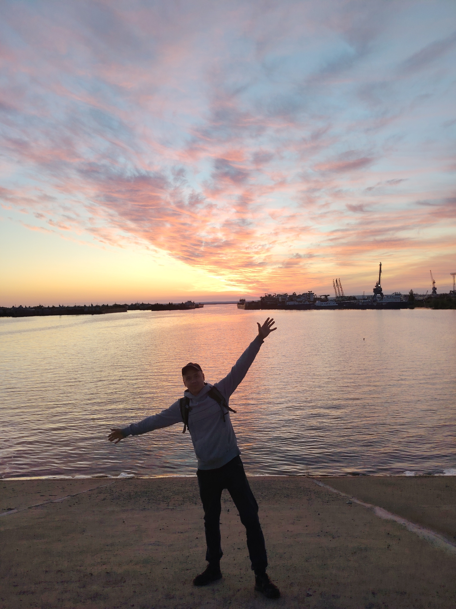 An outdoor photograph captures a person standing on a shoreline at either sunrise or sunset, characterized by a strikingly colorful sky. The horizon glows with warm hues of orange and yellow, transitioning upwards into soft pinks and light blues with wispy cloud formations.
In the foreground, a man is silhouetted against the bright backdrop. He wears a grey hoodie, dark pants, a baseball cap, and a backpack. His arms are outstretched and slightly raised, suggesting a gesture of joy, freedom, or embracing the moment. He stands on a sandy or gravelly bank, with a line or edge visible on the ground beneath his feet.
Beyond the man, a wide body of water, likely a river, stretches across the middle ground, reflecting the soft colors of the sky. In the distance, on the opposite bank, an industrial port area is visible, featuring multiple cranes and several large ships or barges docked along the shore. The low light of dawn or dusk creates a serene and expansive atmosphere. The setting is Chaikovsky, Russia. There is no visible text in the image.