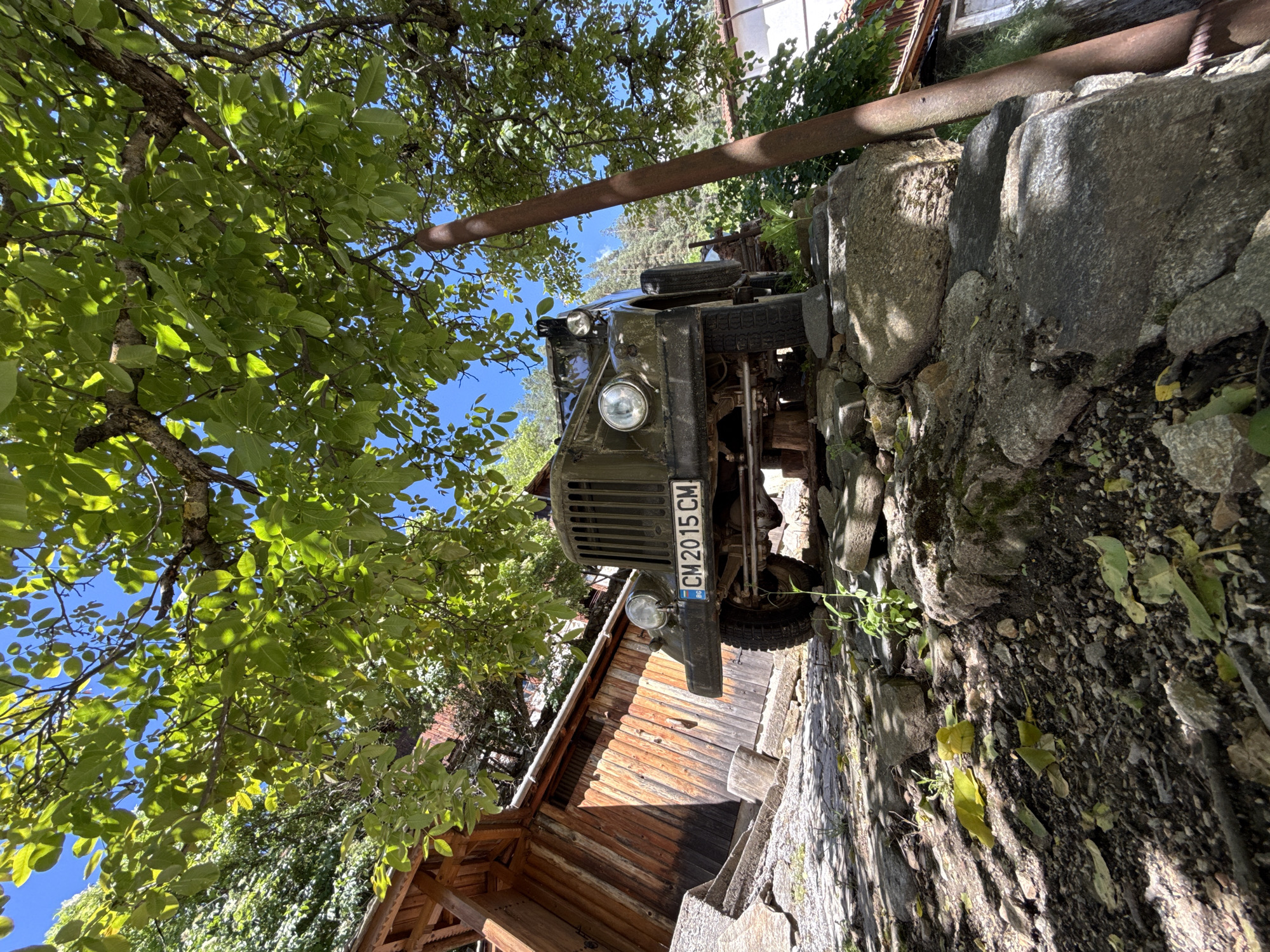 The image captures a low-angle view of a vintage olive-green military truck parked on a rocky incline in Shiroka Laka, Bulgaria. The truck, with a license plate reading "CM 2015 CM," is partially obscured by the lush green canopy of a tree overhead, which casts dappled shadows on the scene. To the left of the truck, a rustic wooden structure with a sloping roof is visible. The ground is a rough terrain of stones, dirt, and fallen leaves. In the background, a tall, rusty metal pole stands vertically to the right of the truck, and hints of houses and trees suggest a rural or mountainous setting. The sky is clear blue.