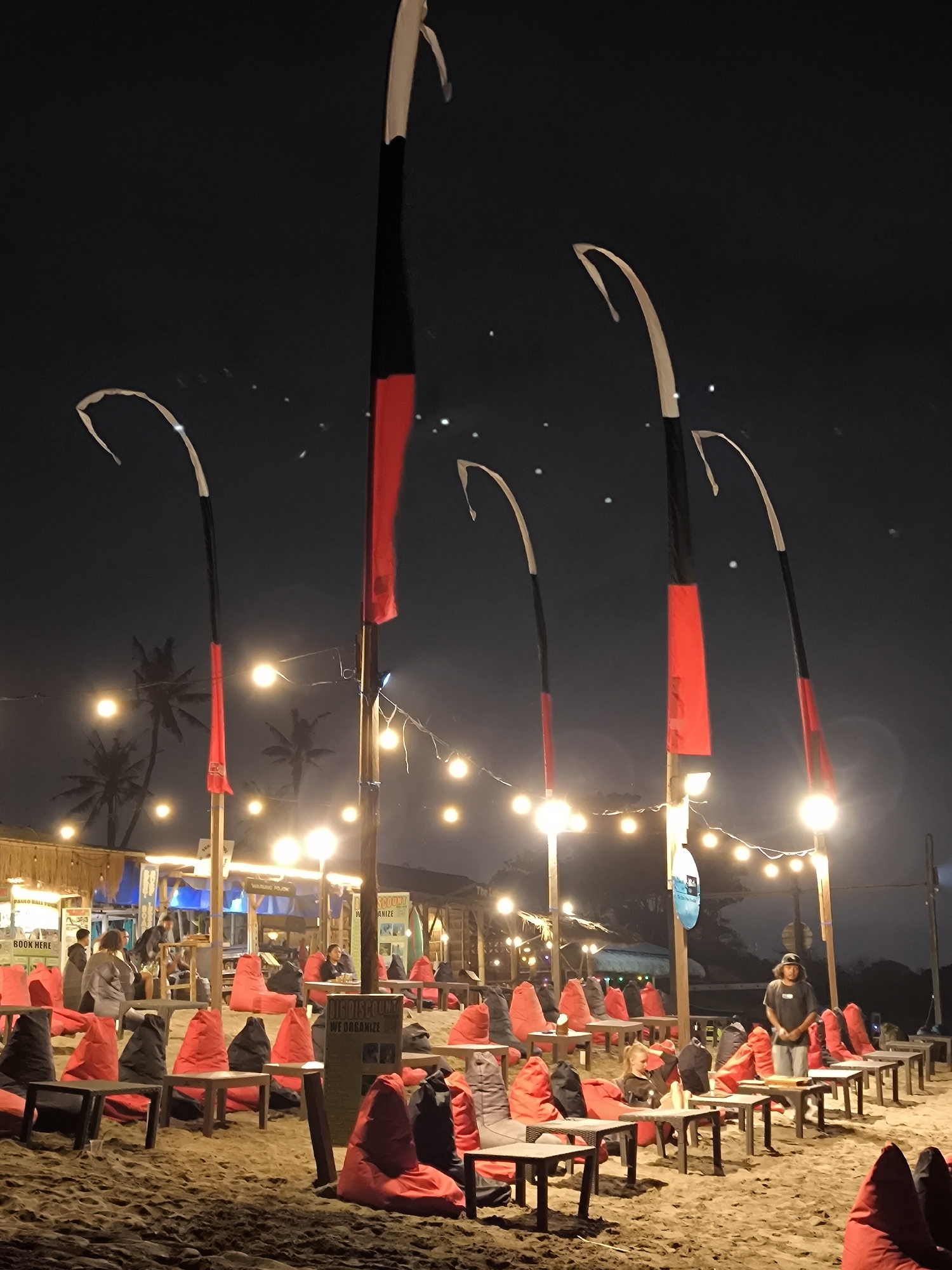 The image depicts an outdoor beach lounge at night in Canggu, Indonesia. Several tall, decorative poles with curved tops are spaced along the sandy beach. Each pole has a segment of red and black fabric hanging from it. String lights are strung between the poles, providing illumination. Numerous small tables are arranged on the sand, surrounded by red and black beanbag chairs. Some people are sitting and standing around the tables, with one person wearing a black t-shirt and cap standing near the front of the seating area. A building or structure is visible in the background, partially obscured by the lighting and people. The sky is dark, suggesting a nighttime setting.