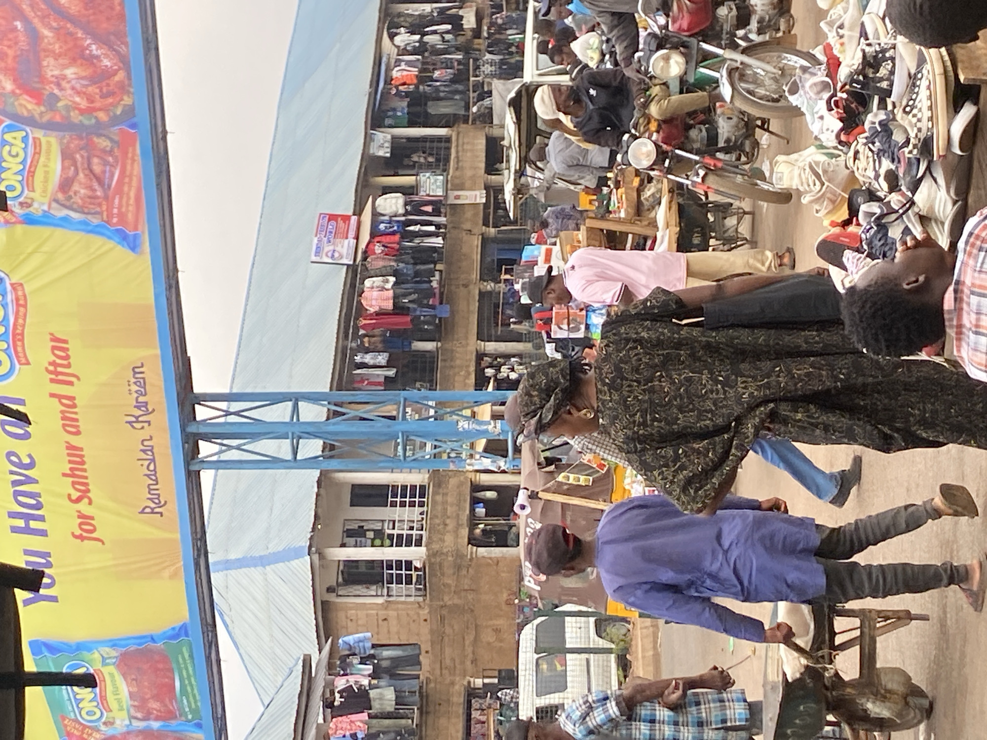 This image depicts a bustling outdoor market scene in Bukuru, Nigeria, likely during the daytime under an overcast sky. The setting includes multi-story buildings lining the street, with market stalls and goods displayed along their lower levels. The ground appears unpaved, adding to the informal market atmosphere.
Numerous people, predominantly men, are visible, engaged in various market activities such as walking, shopping, and manning stalls. In the foreground, a person wearing a patterned top and cap is visible from the back, and another in a blue tunic and dark trousers is operating a hand-powered cart or machine. Several motorcycles, some loaded with goods, are parked or being moved, serving as a common means of transport and commerce. Clothes are seen hanging on racks and piled on tables in front of the buildings, while shoes and other items are displayed on makeshift stalls. A white van is partially visible in the mid-ground.
A large, colorful advertising banner for "ONGA" brand food products dominates the left side of the frame. The banner advertises "Chicken Flavour" and "Beef Flavour" products and prominently displays text such as "You Have an Mama's helping hand for Sahur and Iftar" and "Ramadan Kareem," indicating the photograph was taken during the holy month of Ramadan. Other visible text includes "GREAT TASTE" and "WORLD" on a small sign attached to one of the buildings. The scene conveys the vibrant, daily commercial activity of a local market.
