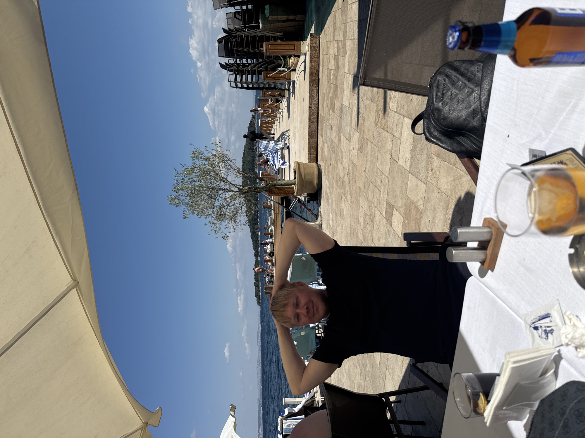 In the city of Prince Islands, Turkey, a man with blonde hair, wearing a black t-shirt, is seen seated at a table outdoors. He has his hands behind his head, and he is looking towards the camera with a slight smile. The table is covered with a white tablecloth and set with drinks and napkins. In the background, there is a body of water, a pier with lounge chairs, and a stacked pile of chairs. The sky is clear and blue.