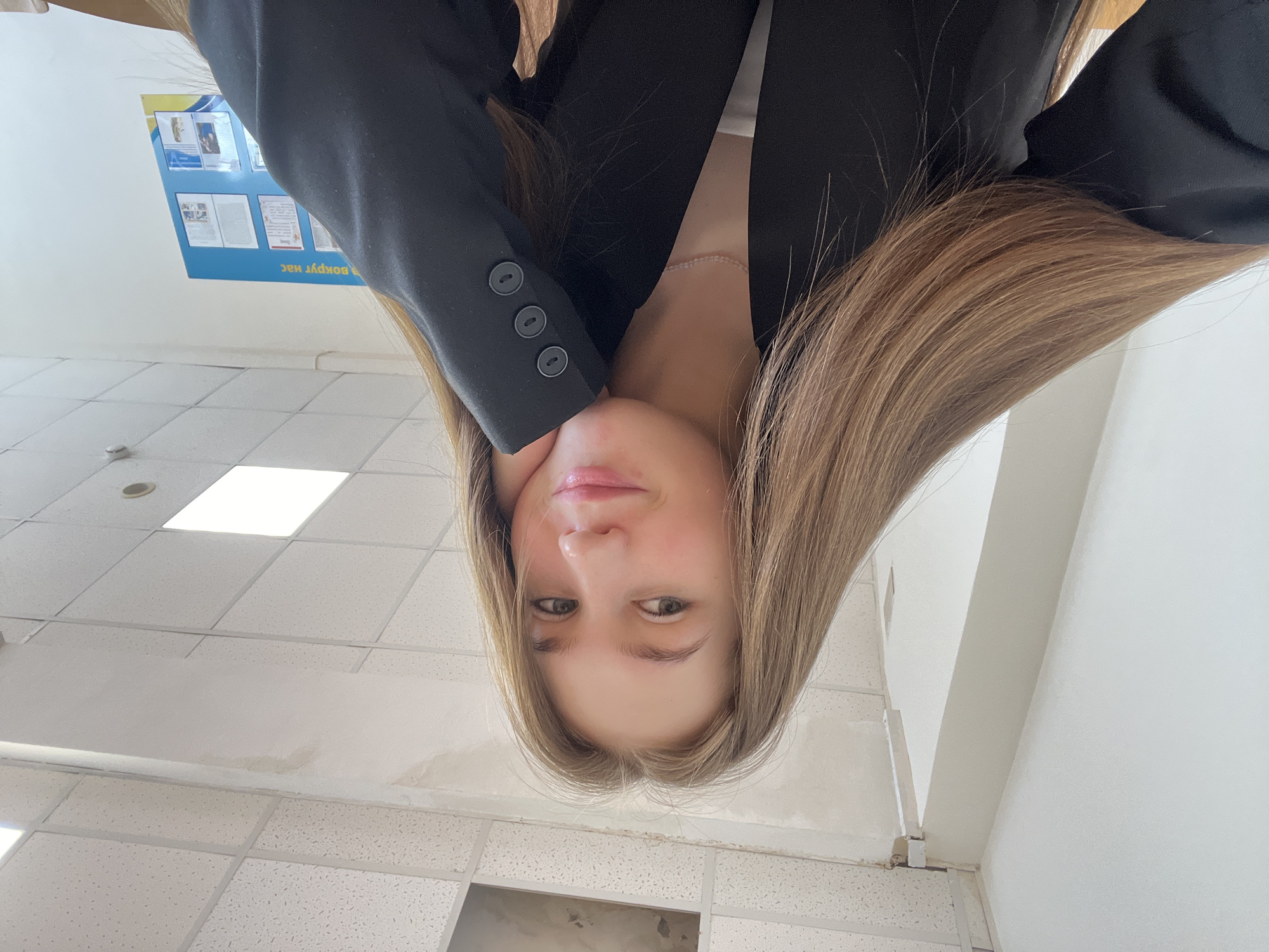 A selfie taken by a woman with long brown hair, wearing a black blazer. The location appears to be indoors, likely a building with a tiled ceiling and walls. A bulletin board with documents is visible in the background. The exact location within Chaikovsky, Russia cannot be determined from the visual information.