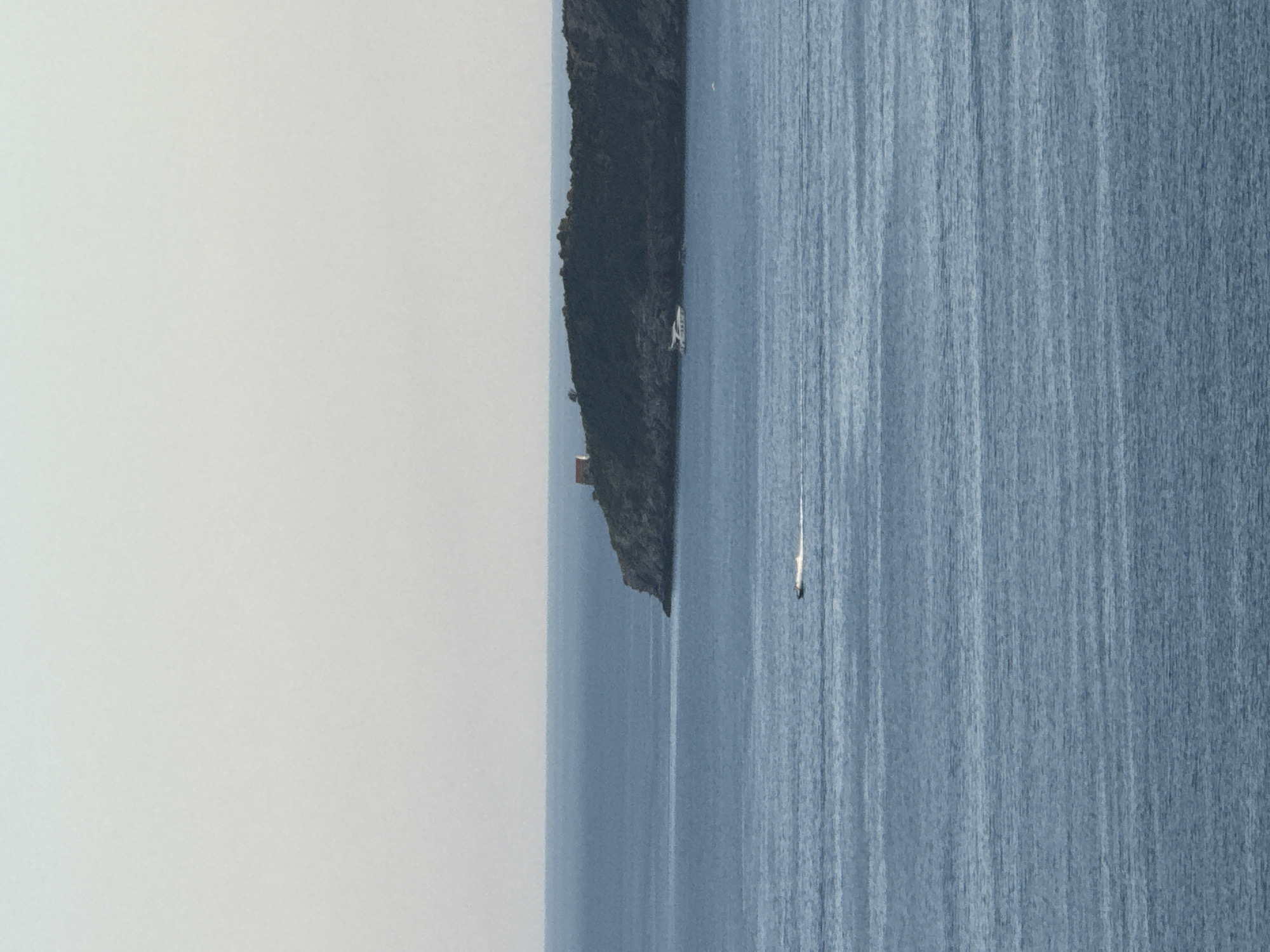 The image shows a body of water with a distant landmass on the horizon. On the landmass, there is a small, red-roofed building situated on a dark, treed area. In the water, two boats are visible. One boat is closer to the shore, appearing larger and white. The other boat is further out, a smaller, dark vessel leaving a white wake. The sky above is a pale, hazy blue. The location is identified as Boreti, Montenegro.