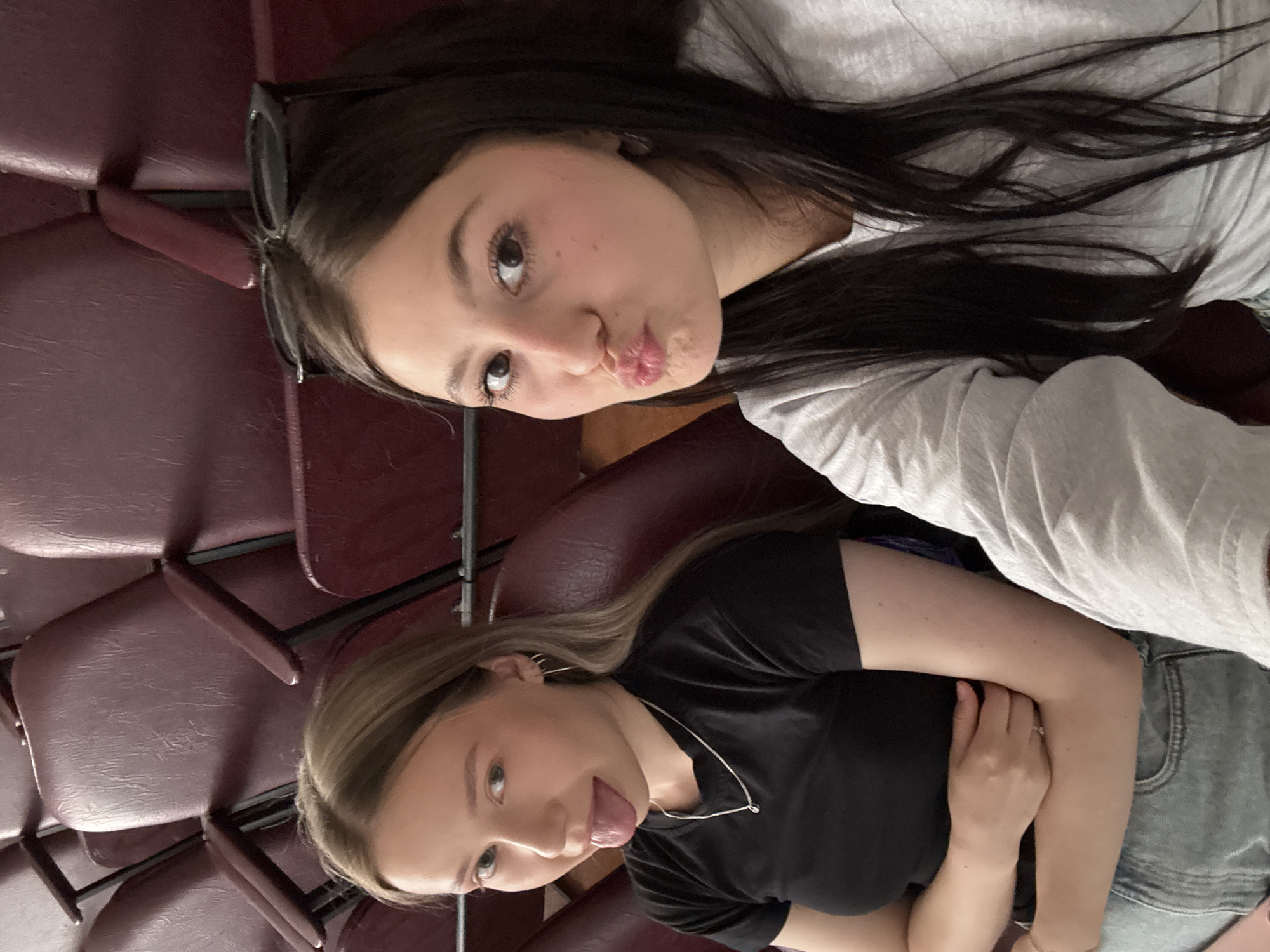 Two young women are sitting in a movie theater in Chaikovsky, Russia. The first woman, with long, dark hair and sunglasses perched on her head, is making a kissing face toward the camera. She is wearing a light gray t-shirt. The second woman, with long, blonde hair, is sticking her tongue out. She is wearing a black t-shirt and blue jeans, and her arms are folded. They are seated in maroon-colored upholstered seats.