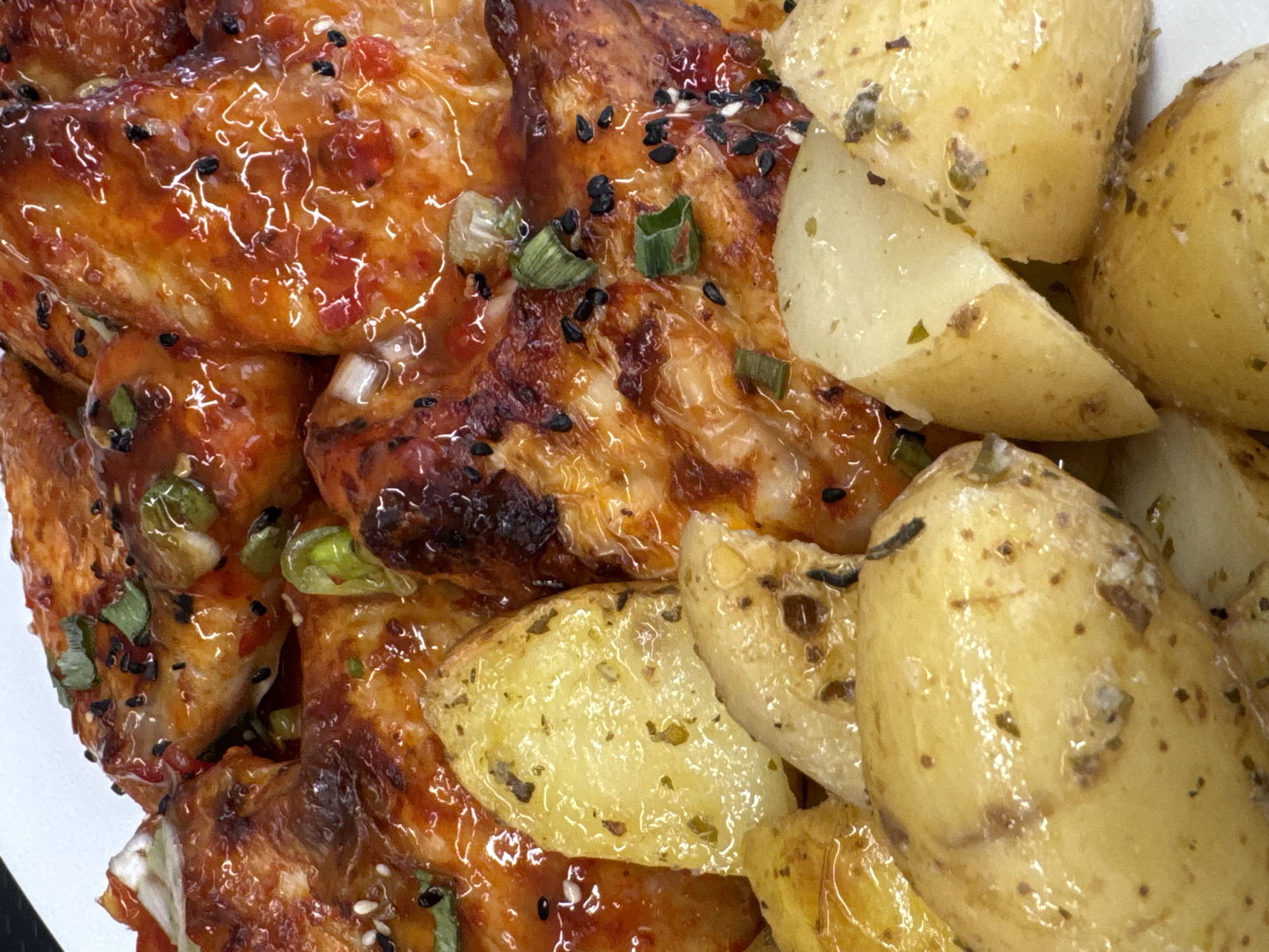 The image is a close-up shot of a plate of food, featuring glazed chicken wings and roasted potato wedges. The chicken wings are coated in a glossy, reddish-orange sauce, sprinkled with black sesame seeds and chopped green onions. The potato wedges are golden brown, appearing to be seasoned with herbs. The food is presented on a white plate, suggesting it is a meal served in a restaurant or at home. The background is out of focus and appears to be a white surface. There is no indication of the location being Athens, Greece.