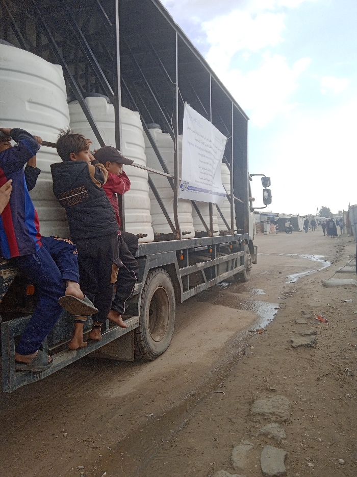 A grey flatbed truck is positioned on a dirt road in what appears to be a camp or informal settlement within Khan Yunis, Palestinian Territory. The truck's bed is loaded with approximately nine large, white, cylindrical plastic water tanks, secured by a dark metal frame. A white banner with text, likely Arabic, and a blue logo is affixed to the side of the truck, facing right. Three young males are riding on the exterior step of the truck on the left side of the frame, holding onto metal bars that support the truck's superstructure. The leftmost child wears a blue and red striped shirt and dark blue pants with green sandals. The middle child wears a dark jacket over dark pants and is barefoot. The rightmost child wears a red jacket and a dark baseball cap, also appearing to be barefoot. The ground beneath the truck is a mixture of dirt and shallow puddles, indicating uneven terrain and recent moisture. Further down the dirt road to the right, various makeshift structures, possibly tents or temporary shelters, line the path, and several figures, some appearing to be women in traditional attire, are visible walking in the distance. The sky is overcast with patches of lighter clouds.