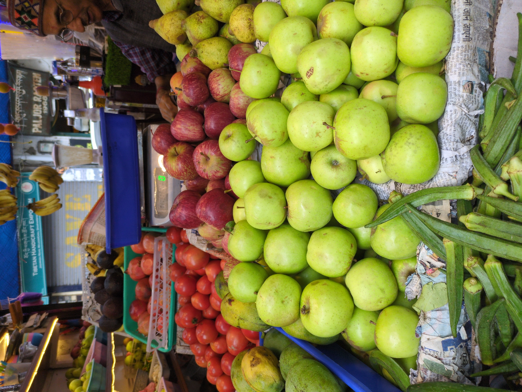 A fruit and vegetable stall is displayed in Dharamshala, India. The stall is packed with various fresh produce including green apples, tomatoes, pomegranates, bananas, and okra. A man wearing a cap and glasses stands behind the counter, looking at the produce. The stall is set up outdoors with shelves in the background holding more fruits and vegetables. The overall scene depicts a vibrant market atmosphere.
