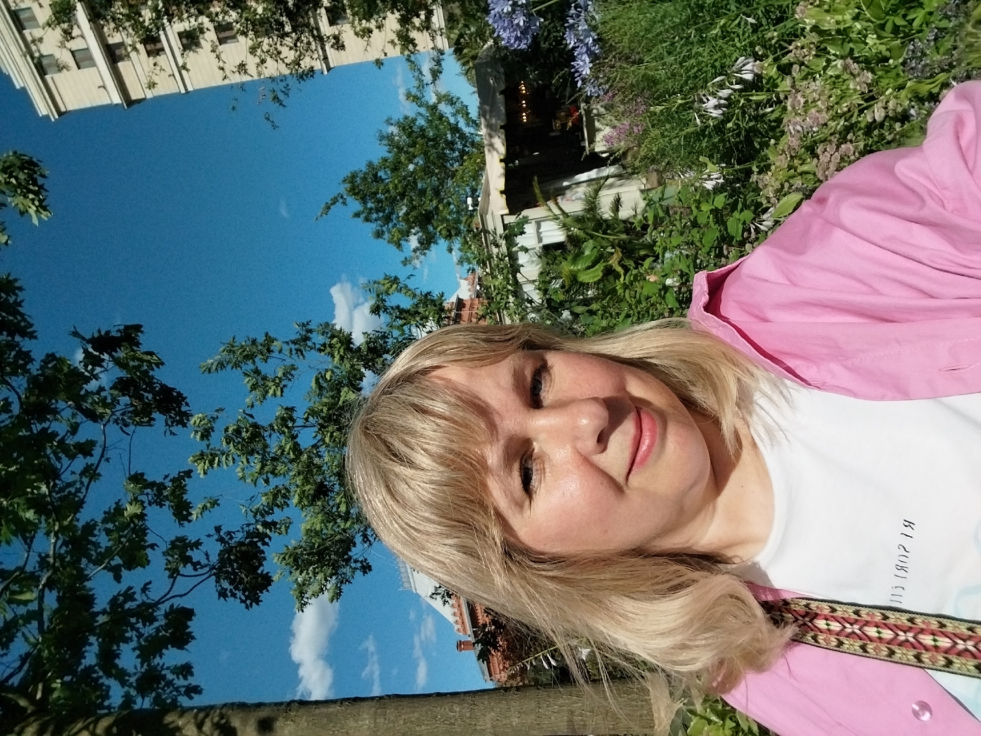 The media file is a close-up, outdoor, selfie-style photograph taken from a low angle. The primary subject is a woman with blonde hair, looking at the camera with a slight smile. She is wearing a bright pink jacket over a white t-shirt. The t-shirt has a decorative embroidered band across the chest.
The background suggests a park or garden setting, with lush green foliage, flowering plants, and trees. In the upper portion of the image, there is a bright blue sky with a few scattered white clouds. Parts of buildings are visible in the background, including what appears to be a cream-colored, multi-story building with visible windows, and hints of red brickwork suggesting older architecture. The image is oriented with the top of the frame tilted.
The lighting indicates it is daytime, likely sunny due to the brightness and the shadows cast by the foliage. The woman's expression conveys a happy and relaxed mood. The text visible on the white t-shirt appears to be in a foreign script, possibly Cyrillic, and is partially obscured. The overall impression is of a pleasant day spent outdoors in an urban or semi-urban environment, possibly Moscow, Russia, given the provided location context.