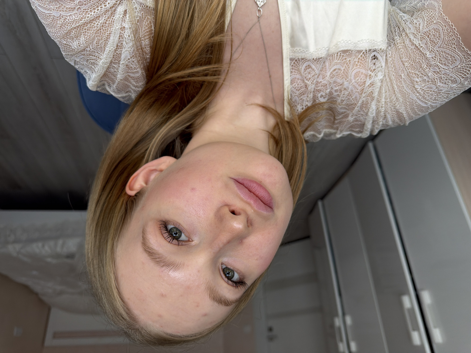 A fair-skinned woman with blonde hair is positioned upside down in the image. She is wearing a white top with lace detailing around the neckline and sleeves, and a delicate necklace with a heart-shaped pendant. The background features a gray wooden floor and white cabinets, suggesting an indoor setting such as a home. The location is Rossiyskiy, Russia.