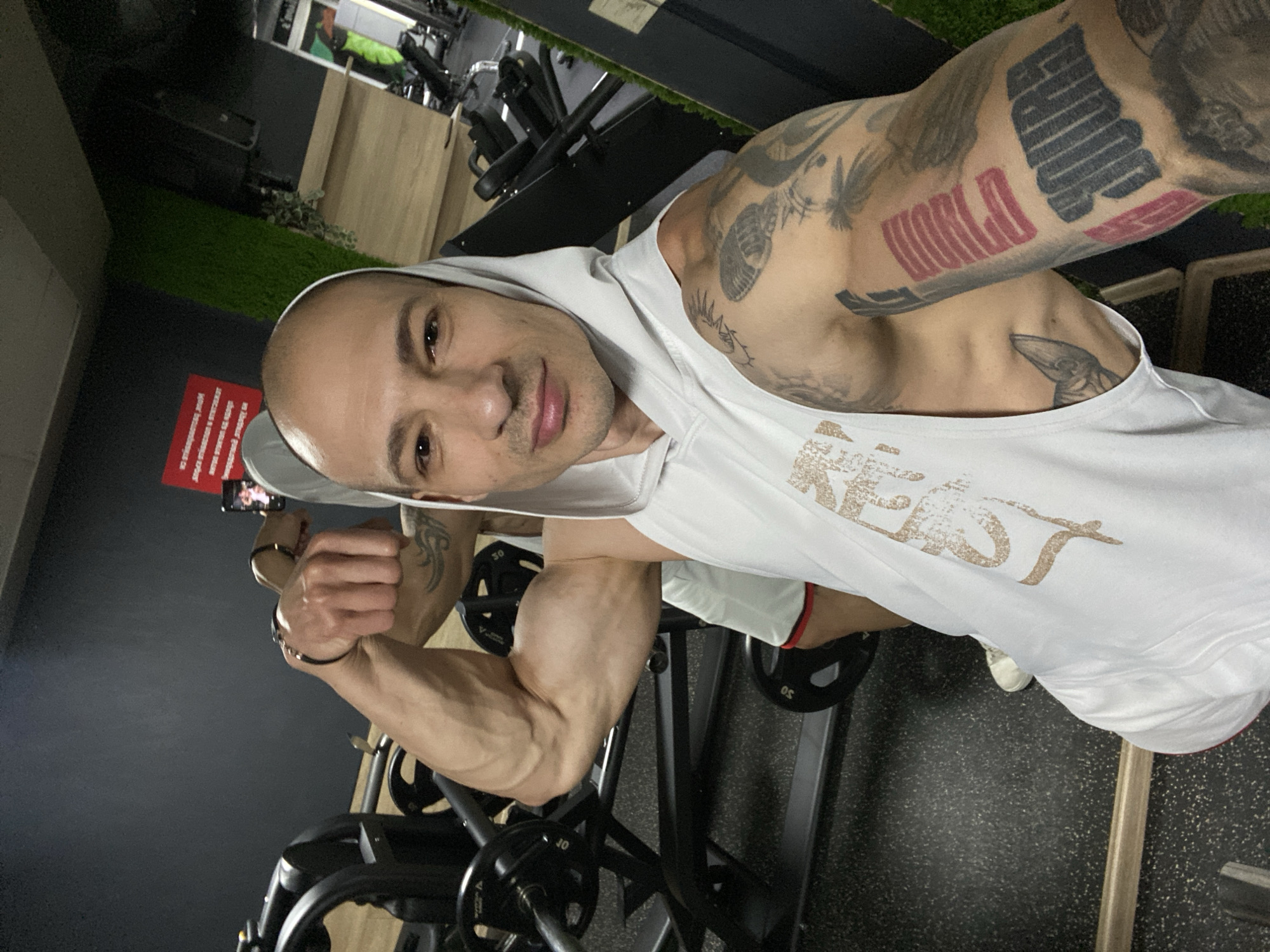 A man is taking a selfie in a gym. He is bald and wearing a white hoodie tank top, white shorts, and white shoes. He is flexing his left arm, which has several tattoos. The gym has weight machines and green Astroturf on the floor. The location is the city of Pleven, Bulgaria.
