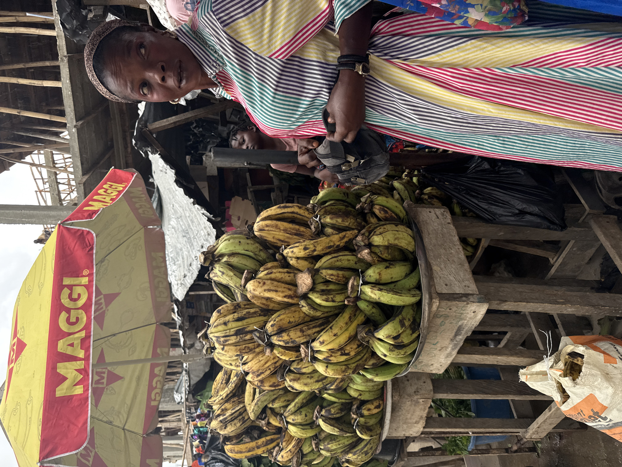 In Ado, Nigeria, a woman stands in a bustling outdoor market under a yellow and red Maggi umbrella. She wears a vibrant, multicolored striped dress and a patterned headscarf, her gaze directed to her right. In her hands, she clutches a small gray bag. Piled high on wooden stands in front of her are numerous bunches of green and yellow bananas, some with brown blemishes. Behind her, a younger person, possibly a child, is visible in the background, partially obscured by wooden structures and market stalls. The scene is characterized by the abundance of produce and the lively atmosphere typical of a Nigerian market.