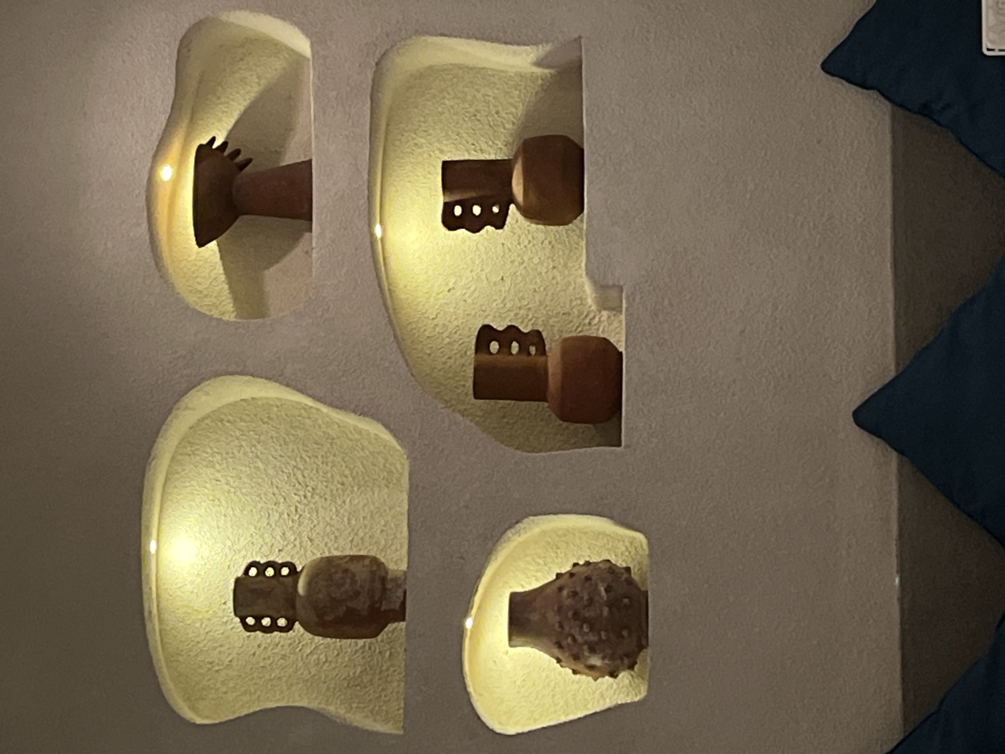 The image shows four recessed wall light fixtures arranged in two columns and two rows on a textured gray wall. The fixtures are a light beige color and have curved, organic shapes. Each fixture houses a brown decorative element. From top left to bottom right, the elements appear to be: a stylized hand; a stylized key-like shape with a knob and a series of holes; a second stylized key-like shape with a knob and a series of holes; and a textured spherical object. The light source within each fixture illuminates the decorative element and casts a glow around the fixture. The location is Unknown, Indonesia. A portion of two dark blue throw pillows are visible on the right side of the frame, near the right edge.