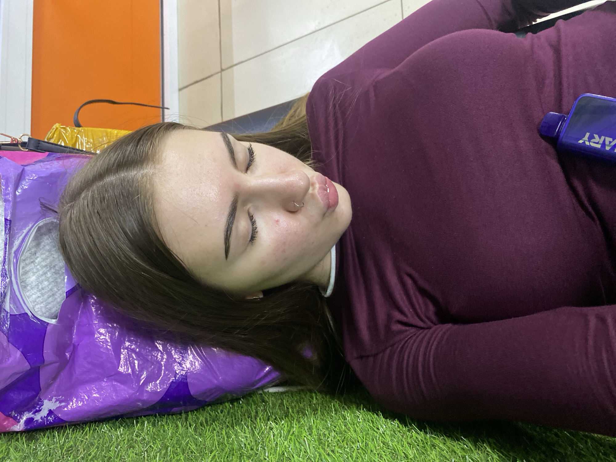 A young woman is lying down on a purple mat with green artificial grass to her left. She is wearing a dark maroon long-sleeved shirt. Her eyes are closed and her lips are pursed. A small object, possibly a vape, is resting on her chest. In the background, there is an orange surface and some beige tiling. The scene appears to be indoors, possibly in a commercial or recreational space.