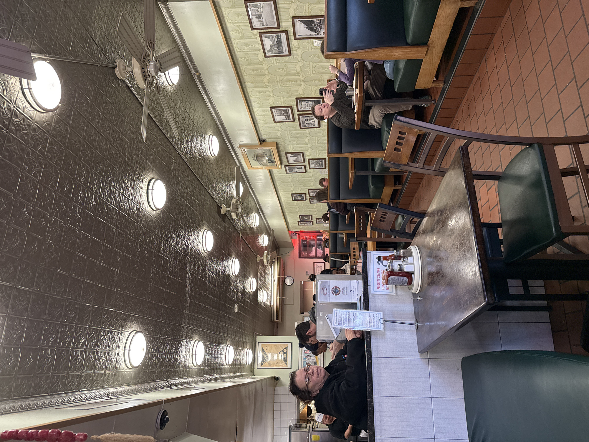 The image shows the interior of a dining establishment, likely a diner or restaurant, possibly in New York City. The walls are covered in a pressed tin pattern, punctuated by round sconces. Ceiling fans are interspersed along the length of the room. The seating consists of booths along one side and tables with chairs on the other. People are seated at the tables, some of whom are looking at their phones. Framed pictures adorn the wall above the booths. In the foreground, a person is looking at the menu while standing at the counter, behind which is a service area and the restaurant continues further back in the frame, seemingly full of patrons. The floor appears to be tiled. A table with condiments sits in the foreground.