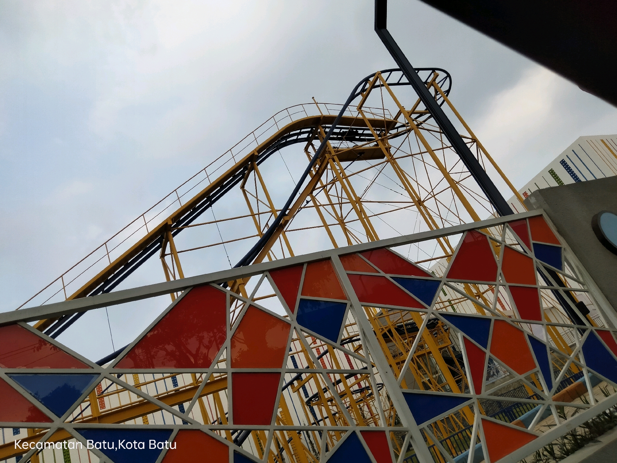 An outdoor perspective captures a section of a yellow roller coaster structure against an overcast sky. The roller coaster track, with its black railing, curves upwards and to the left. A large, black metal pole is visible on the right, supporting part of the structure. In the foreground, a decorative metal frame is adorned with triangular panels of red, orange, and blue. Behind this frame, the intricate yellow supports of the roller coaster are visible. To the far right, a portion of a white building with colored vertical stripes can be seen. Text in the lower left corner reads "Kecamatan Batu, Kota Batu," indicating the location as Batu District, Batu City, Indonesia. No people are visible in the image.
