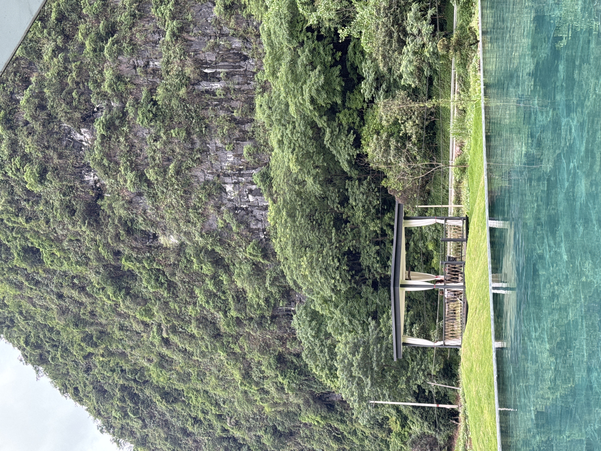 The image depicts a serene scene in Guilin, China, featuring a pool of clear water reflecting the surrounding landscape. Adjacent to the pool is a verdant lawn with a gazebo structure. The backdrop is dominated by a steep, rocky mountain covered with lush greenery. The gazebo appears to be an area for relaxation, possibly for guests at a resort. The location is idyllic, emphasizing Guilin's natural beauty.