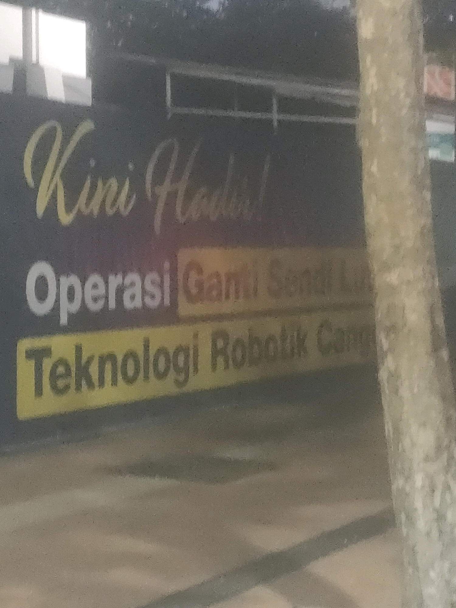 The image shows a sign with text in Indonesian, indicating a location in Surabaya, Indonesia. The sign is dark with yellow and white lettering. The top line of text, in a stylized yellow font, reads "Kini Hadir!". Below this, a line of white text says "Operasi Ganti L". A third line, in yellow, reads "Teknologi Robotik Can". The background is dark, with some blurred light reflections at the top left, possibly from windows. A textured, light-colored tree trunk is visible on the right side of the frame, obscuring part of the background. The ground in front of the sign is a neutral, light brown. The overall impression is of an outdoor sign, likely advertising a business or event.