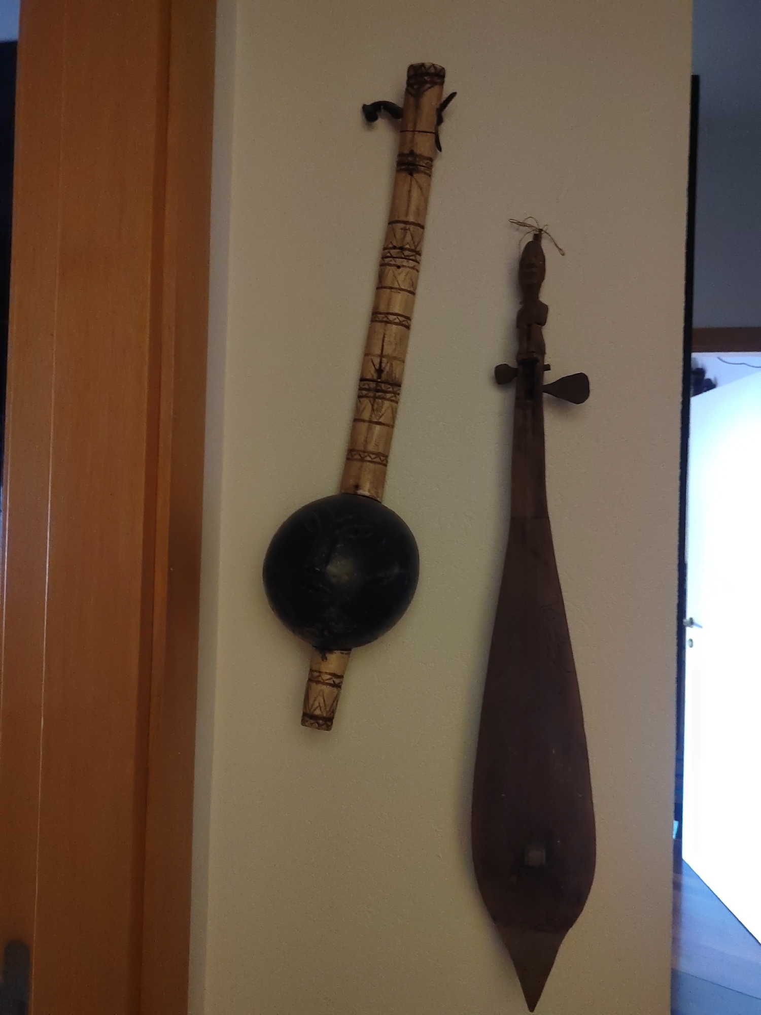 Two African musical instruments are hanging on a cream-colored wall, which appears to be an interior wall of a room. To the left of the frame, a portion of a wooden door or frame is visible. The wall exhibits a fine texture.
The instrument on the left is a stringed instrument with a long, cylindrical neck made of bamboo or similar material. The neck is decorated with intricate geometric patterns carved or burned into the surface. At the lower end of the neck, a large, round, black gourd serves as the resonating body. A single string is visible attached to the top of the neck and extending downwards, presumably to the gourd or a bridge not clearly visible. This instrument appears to be a kora or a similar lute-like instrument.
The instrument on the right is also a stringed instrument, but it has a different construction. It features a narrow, elongated body made of dark wood, tapering to a point at the bottom. The top of the instrument has a carved wooden head, possibly depicting a human or animal figure. Two small, wing-like projections extend from the sides of the neck. A single string is attached to the top of the head and stretches downwards along the body. This instrument resembles a goje or a similar bowed lute.
The lighting in the scene is somewhat dim, with a brighter light source coming from the right side of the frame, possibly from a window or an open doorway, casting some light onto the right side of the wall and the instrument. The overall impression is that these are decorative items displayed in a home or a cultural space. The image does not contain any explicit geographical markers that would place it in Porto Recanati, Italy.